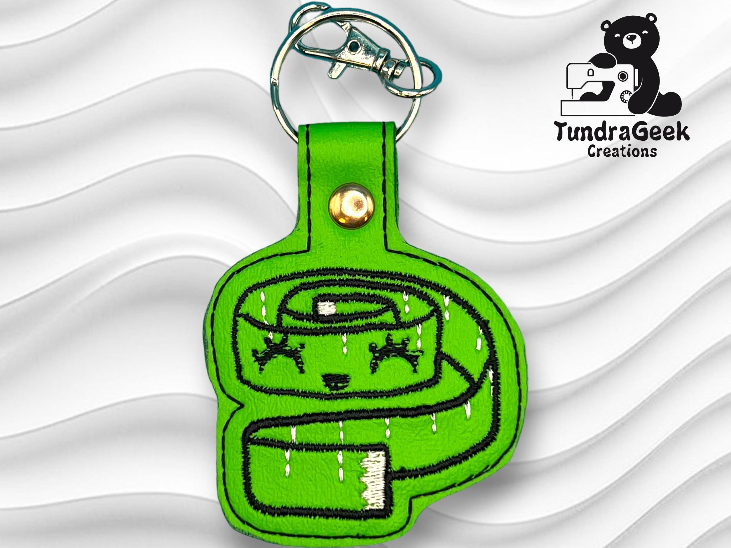 A green embroidered keychain shaped like a coiled snake with a cute face, attached to a metal key ring, displayed on a white textured background.