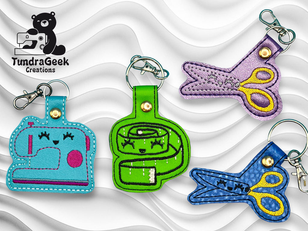 Four embroidered keychains shaped like a sewing machine, tape measure, and two pairs of scissors are displayed on a wavy white background, with the TundraGeek Creations logo in the corner.