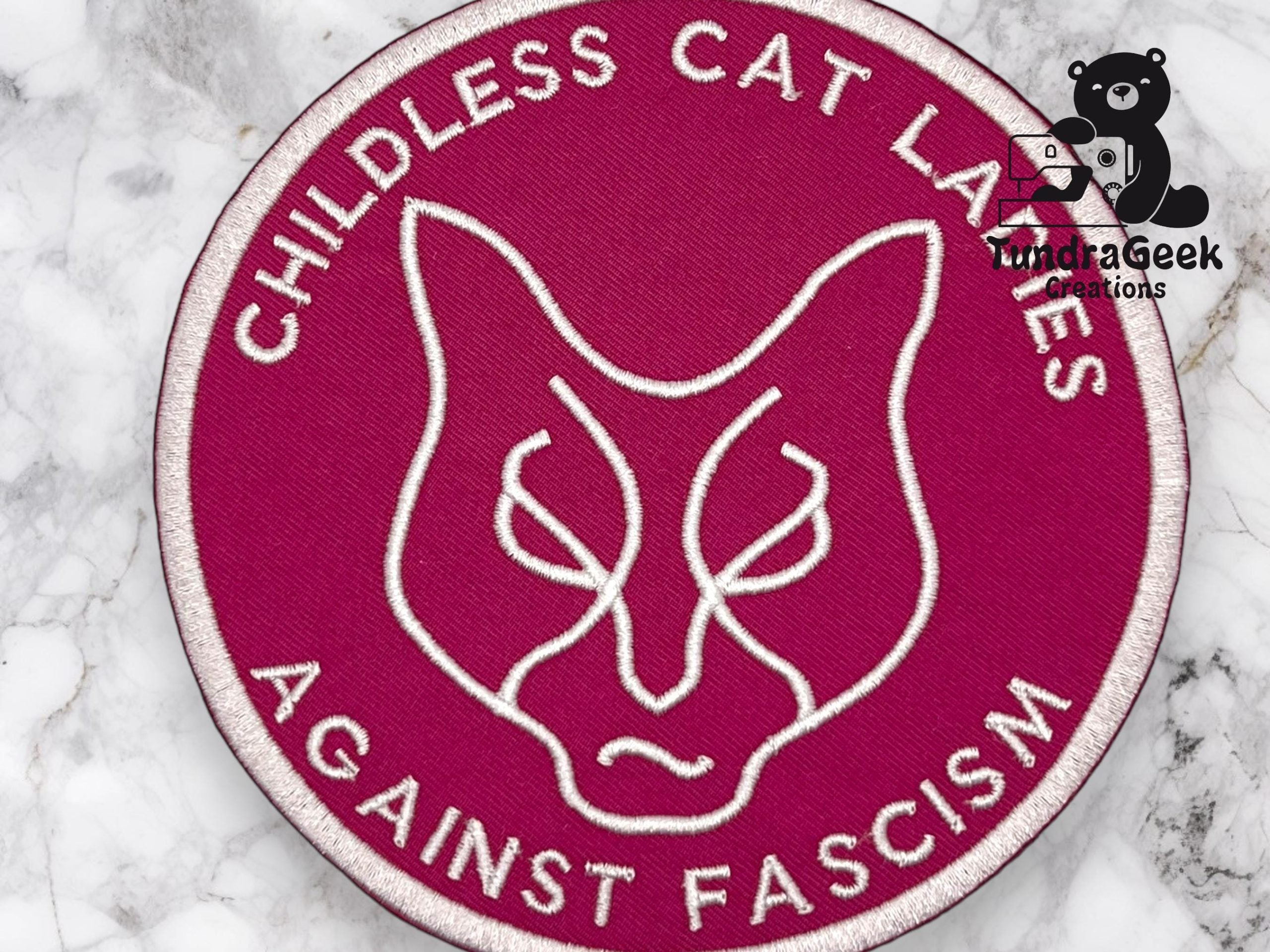 Round patch with a stylized cat face and the words CHILDLESS CAT LADIES AGAINST FASCISM embroidered in white on a magenta background, placed on a marble surface.