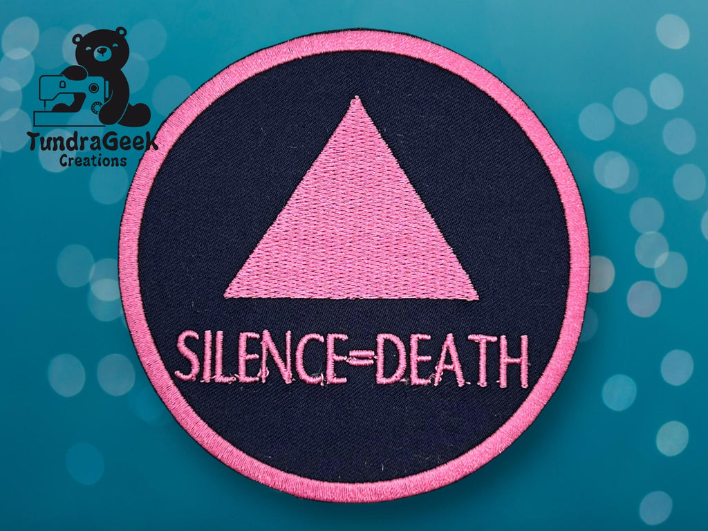 A circular patch with a pink triangle above the words SILENCE = DEATH on a black background, bordered in pink, displayed on a blue background.