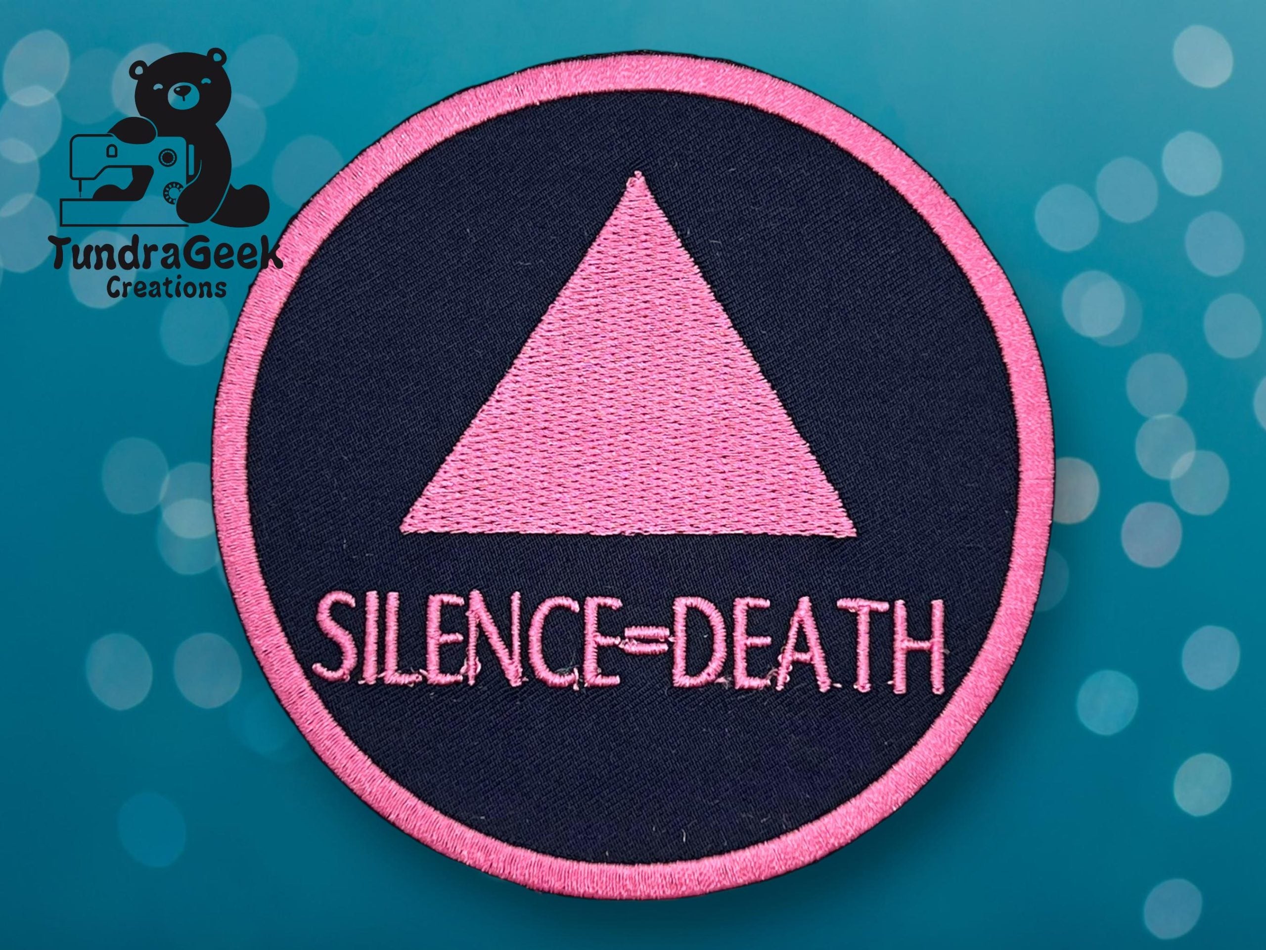 A circular patch with a pink triangle and the words SILENCE=DEATH on a black background, bordered in pink, against a teal backdrop.