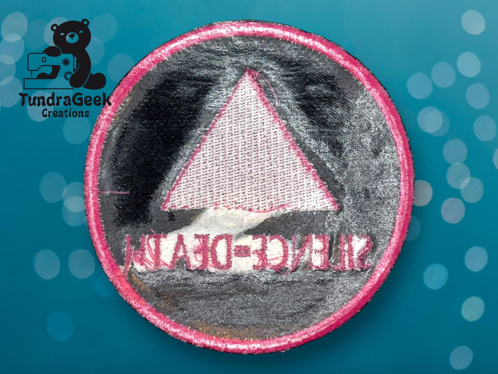 A round embroidered patch with a pink border, featuring a pink triangle and mirrored text reading “SILENCE=DEATH,” on a blue background with bokeh effect and a TundraGeek Creations logo.