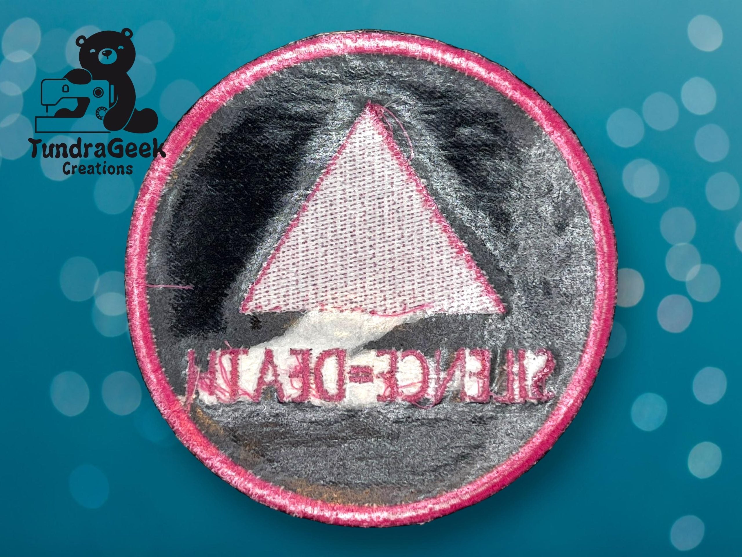 A round embroidered patch with a pink border, featuring a pink triangle and mirrored text reading “SILENCE=DEATH,” on a blue background with bokeh effect and a TundraGeek Creations logo.