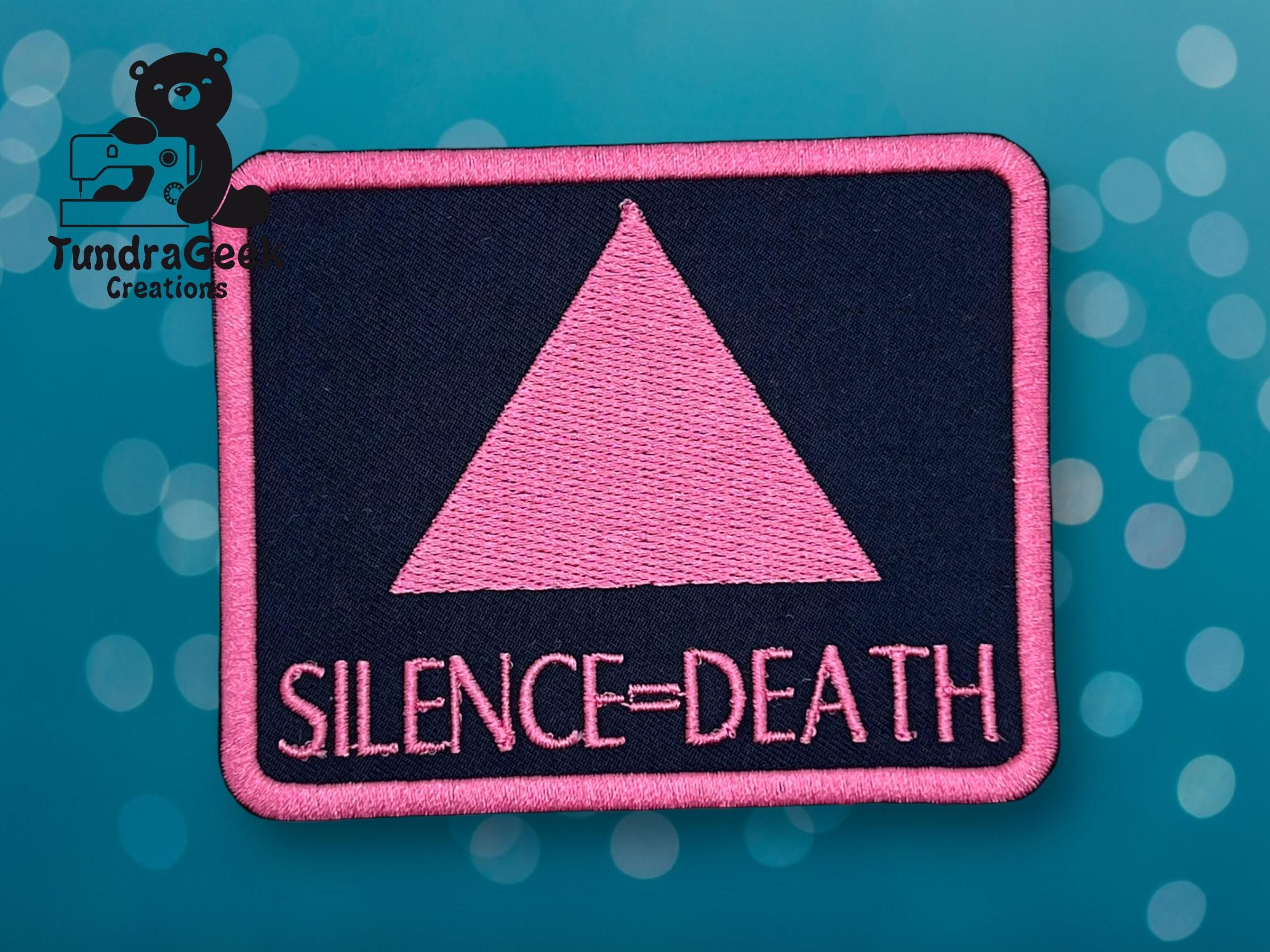 A rectangular patch with a pink triangle above the words SILENCE = DEATH on a dark background, with a bokeh effect and a bear logo in the upper left corner.