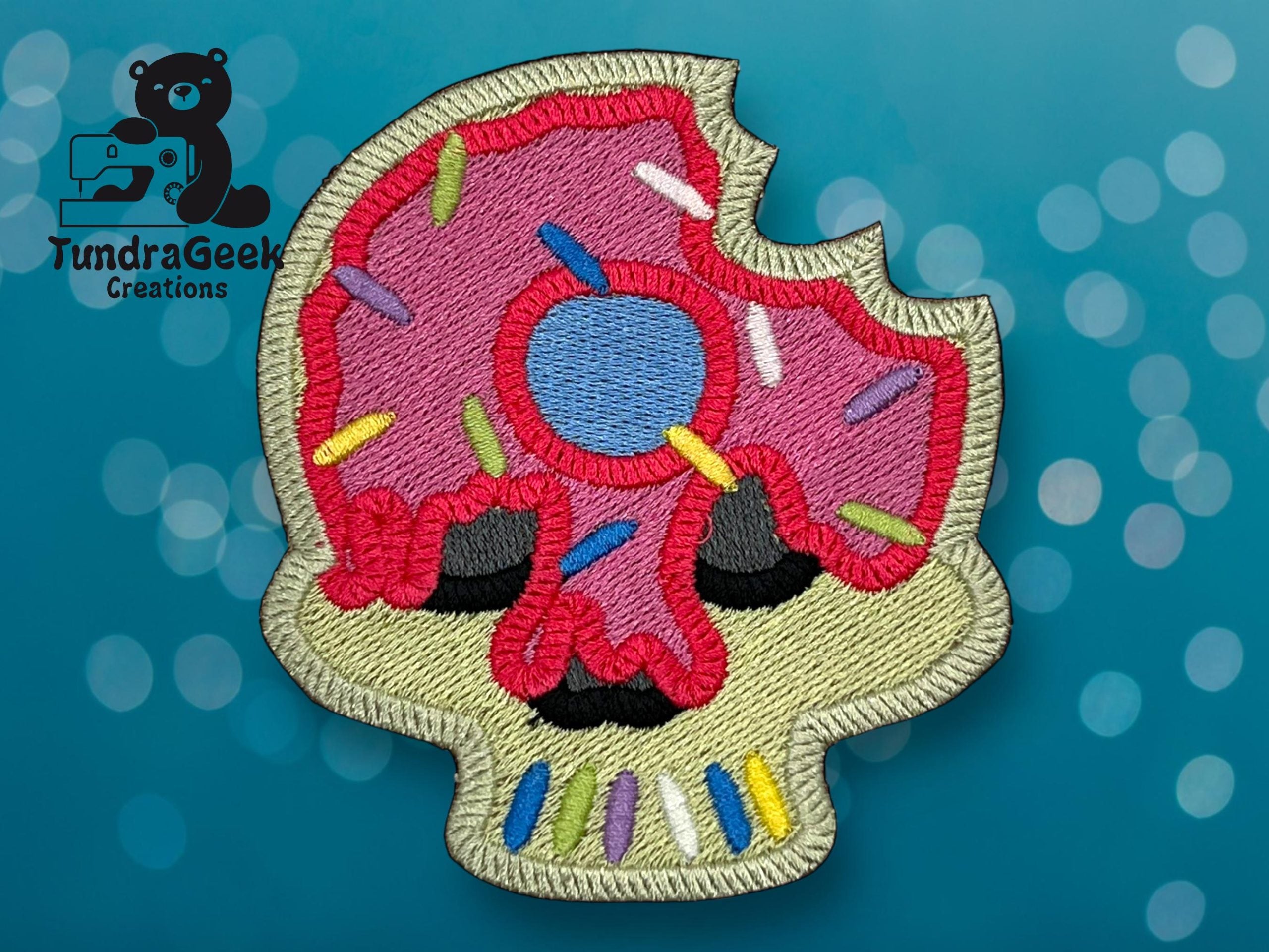 Embroidered patch of a skull with pink icing, sprinkles, and a bite taken out, set against a blue background with circular light patterns.