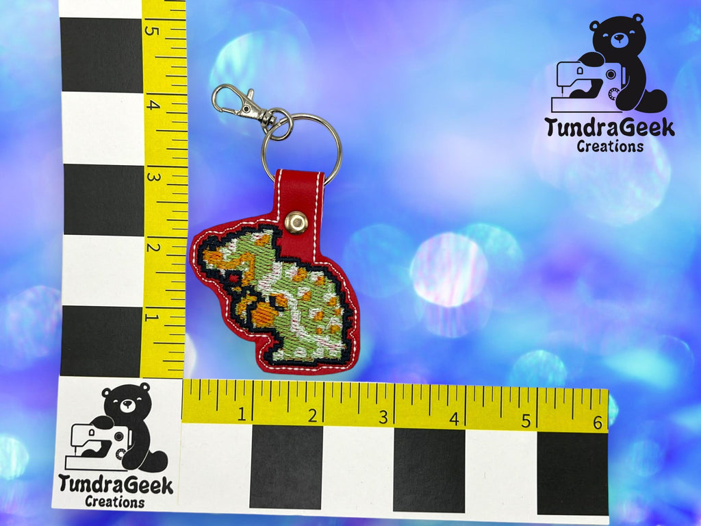 A small keychain with a cross-stitched dinosaur design on red backing is shown next to measuring rulers for scale, with a TundraGeek Creations logo in the background.