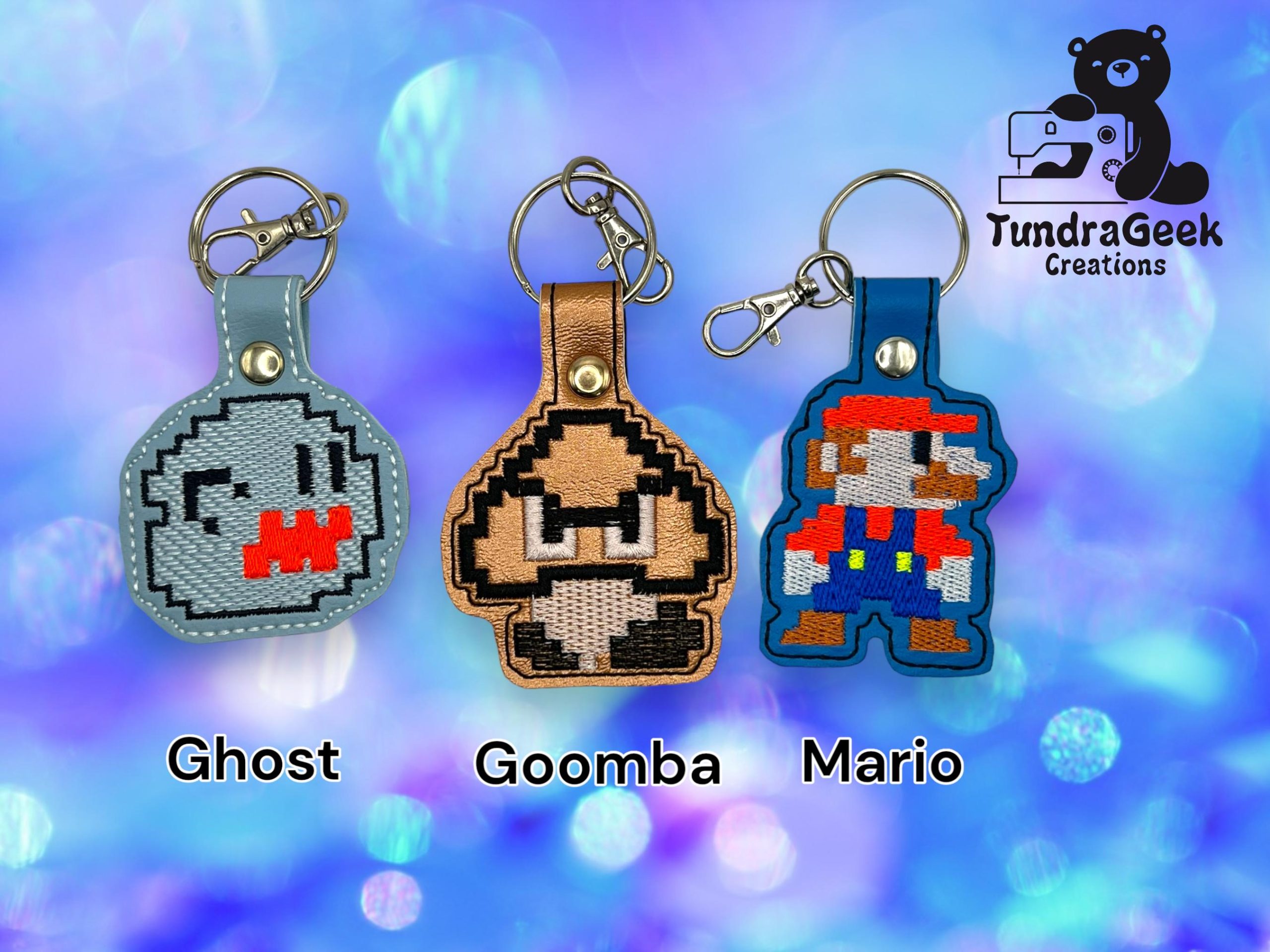 Three pixel art keychains of Ghost, Goomba, and Mario characters are displayed against a blue bokeh background, with the TundraGeek Creations logo in the top right corner.