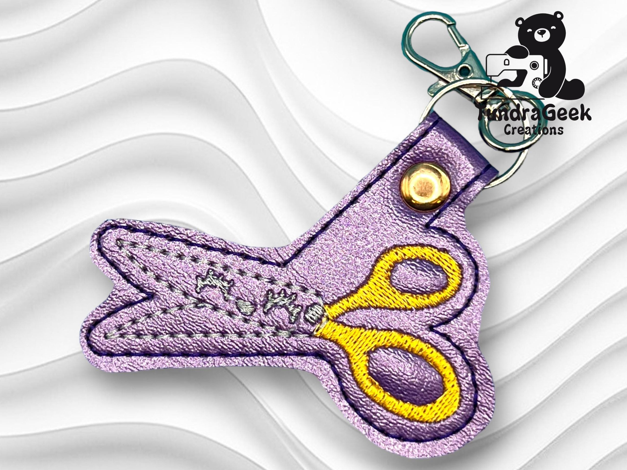 A keychain in the shape of scissors, made of purple and yellow fabric with embroidered details, attached to a clasp.