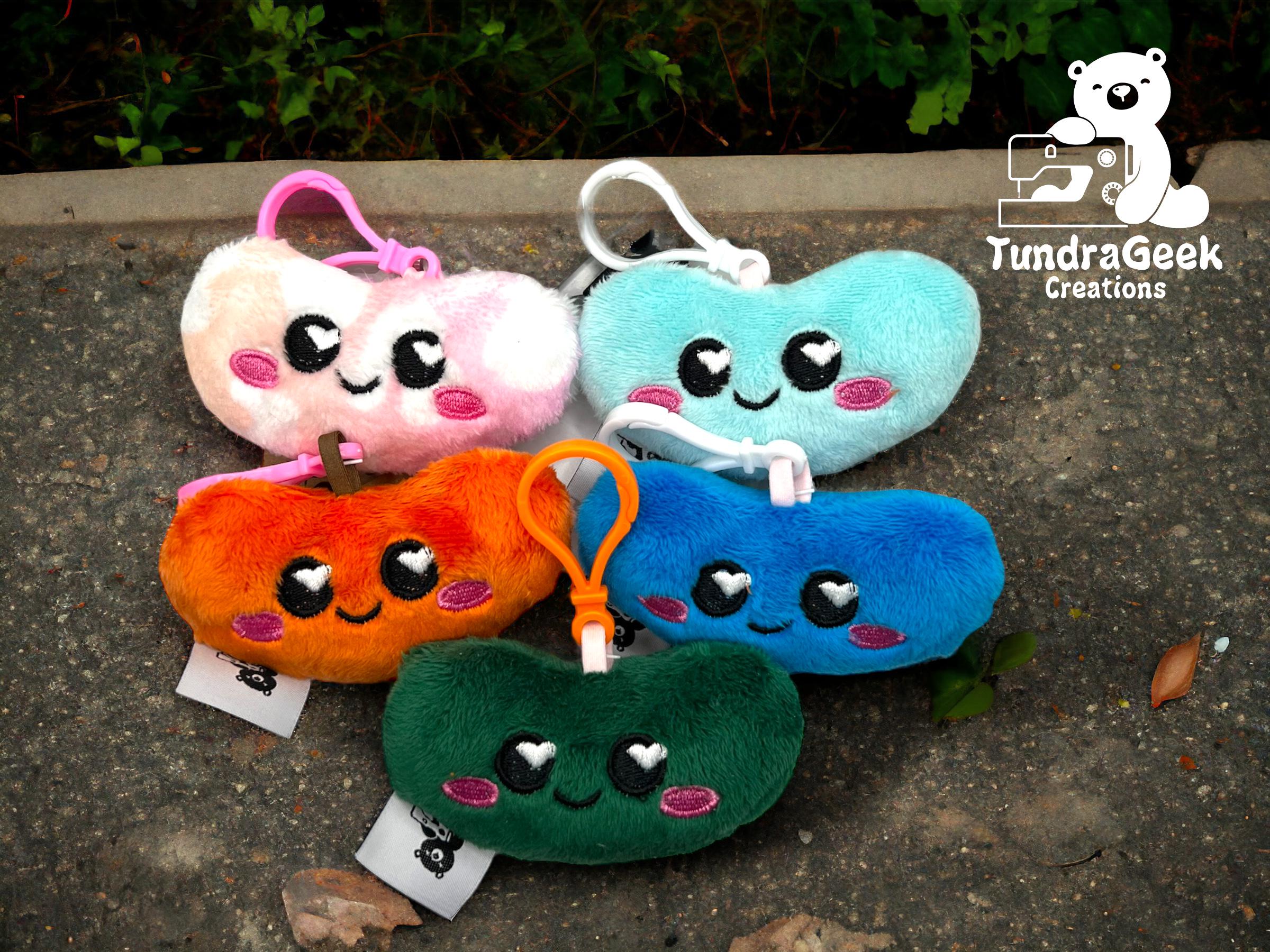 Six colorful, plush keychains shaped like smiling beans are arranged on a gray pavement, with “TundraGeek Creations” and a bear logo in the top right corner.