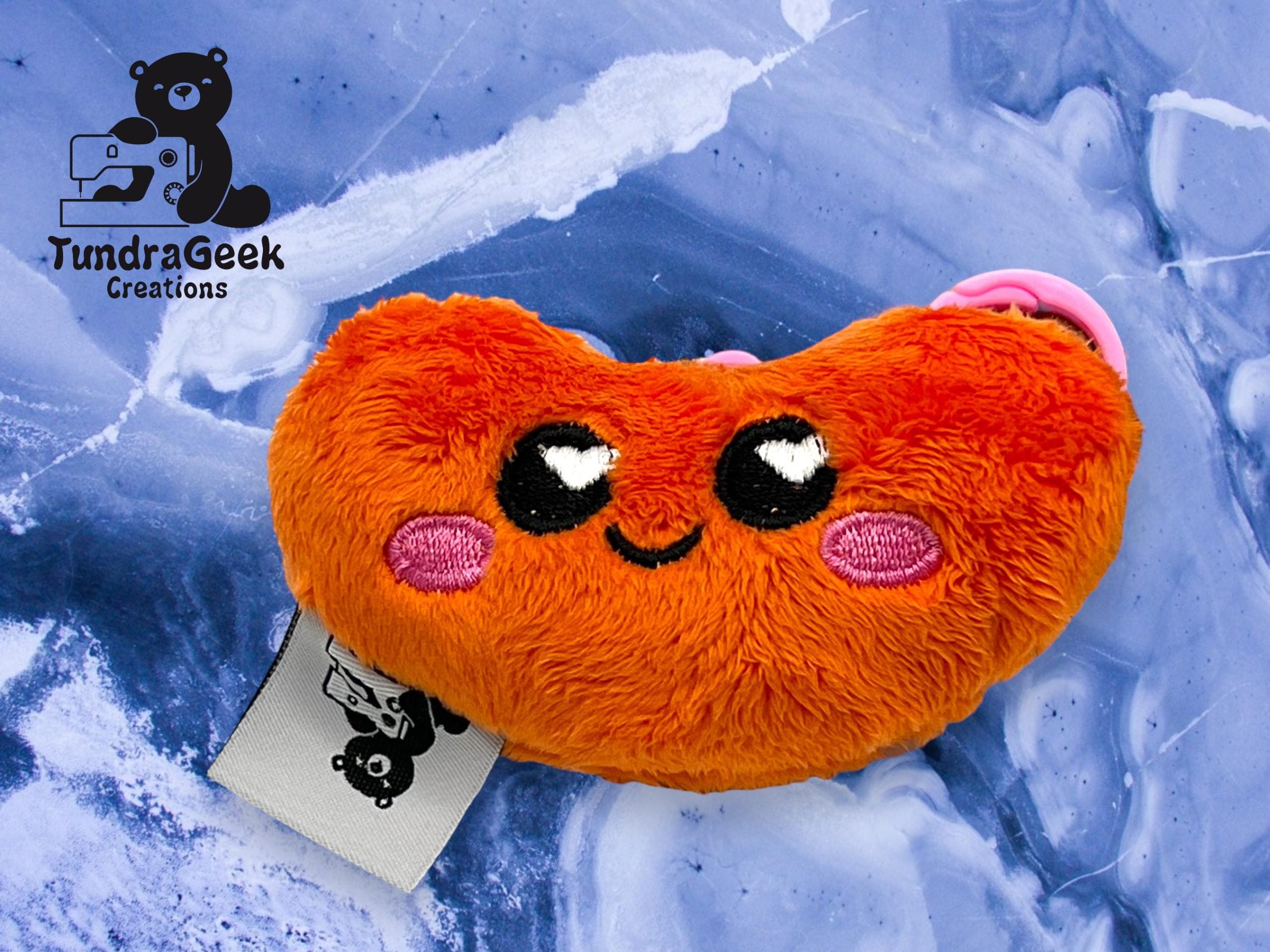An orange plush toy shaped like a bean with embroidered eyes, cheeks, and a smiling mouth, placed on a blue marbled background. TundraGeek Creations logo is visible in the corner.