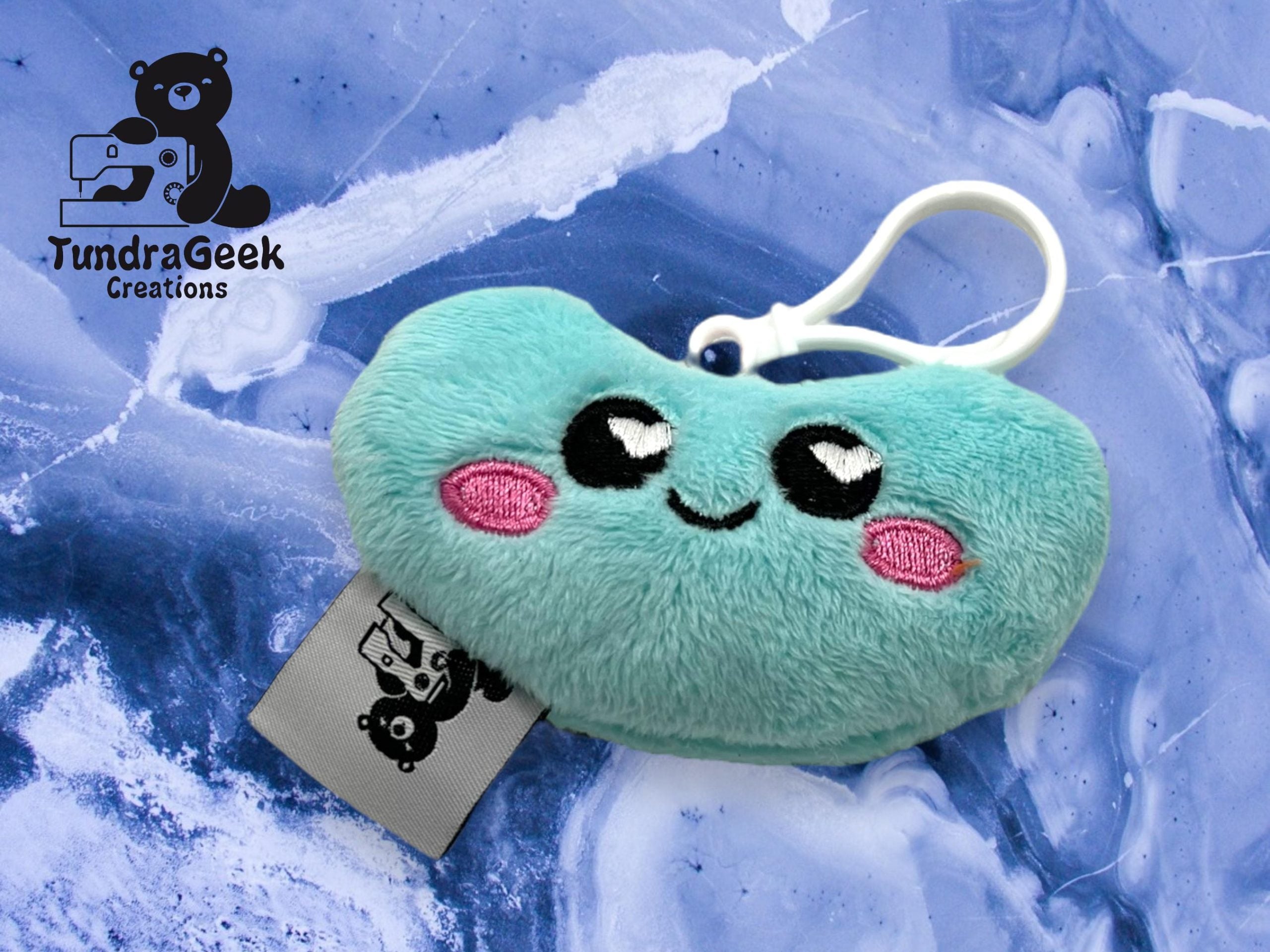 A plush keychain shaped like a smiling teal bean with pink cheeks, displayed on a blue marbled background with the TundraGeek Creations logo in the corner.