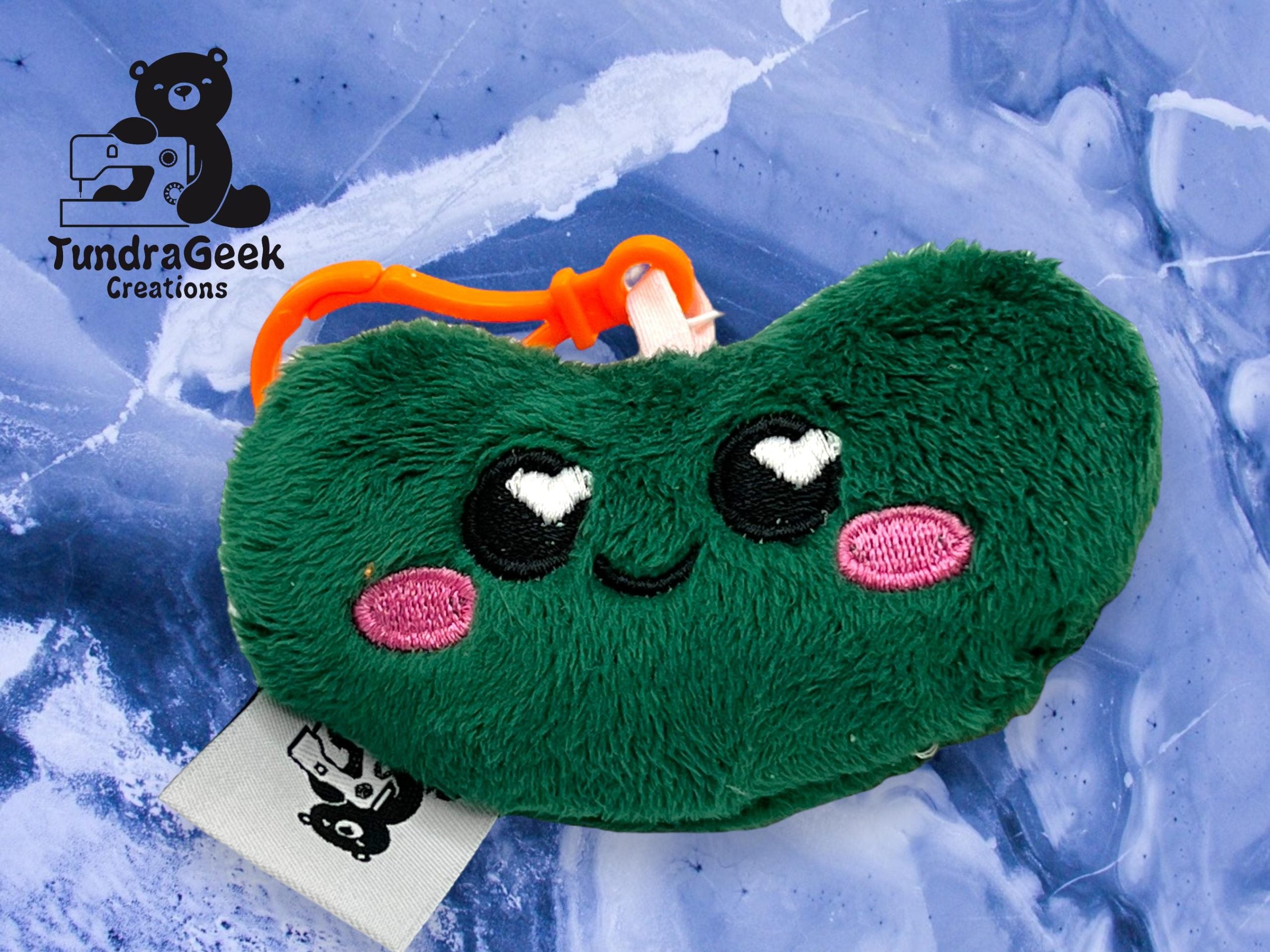 A green plush keychain shaped like a bean with embroidered eyes and pink cheeks, placed on a blue marbled background. The TundraGeek Creations logo is in the top left corner.