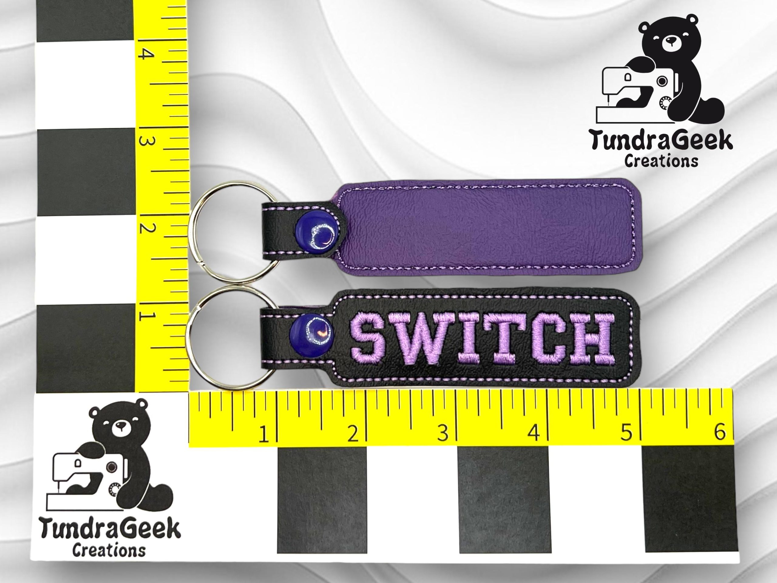 Two rectangular keychains, one plain purple and one purple with SWITCH embroidered in white, shown on a yellow and black measurement grid. TundraGeek Creations logo visible.