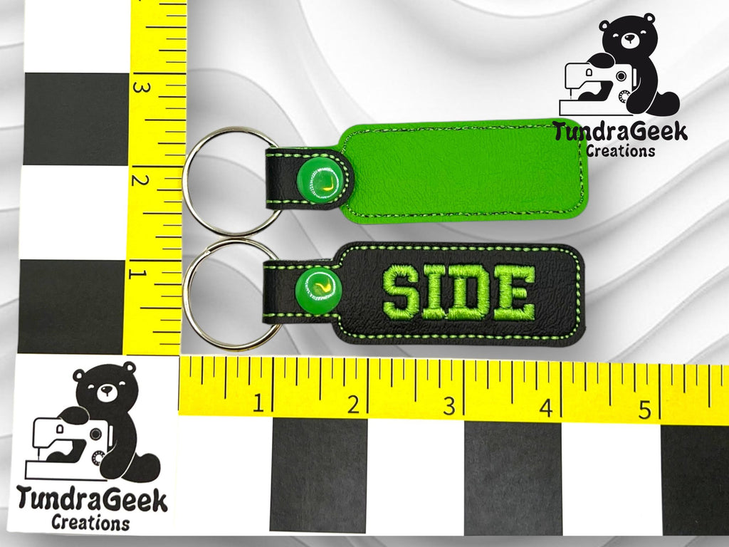 Two green and black keychains, one blank, one with SIDE embroidered, are placed on a ruler background with TundraGeek Creations bear logo.