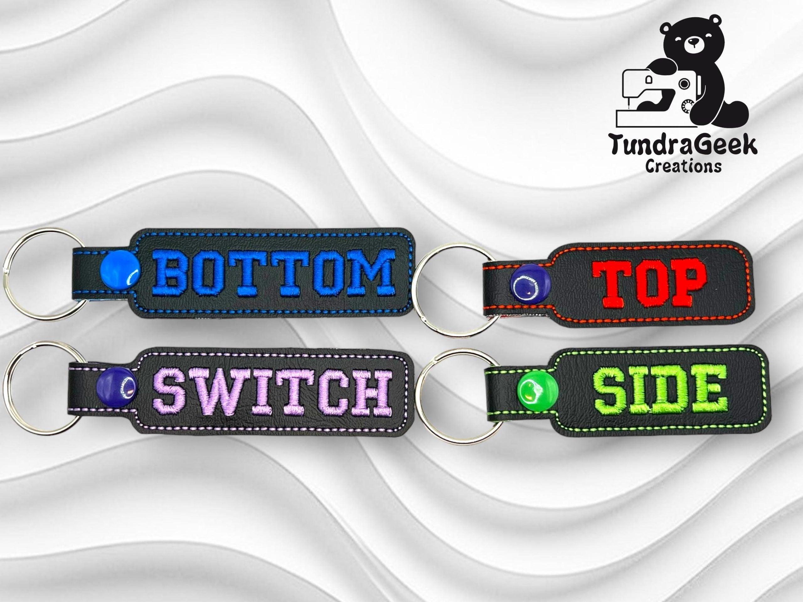 Four embroidered keychains labeled BOTTOM, TOP, SWITCH, and SIDE in different colors on a wavy white background. TundraGeek Creations logo in the top right corner.