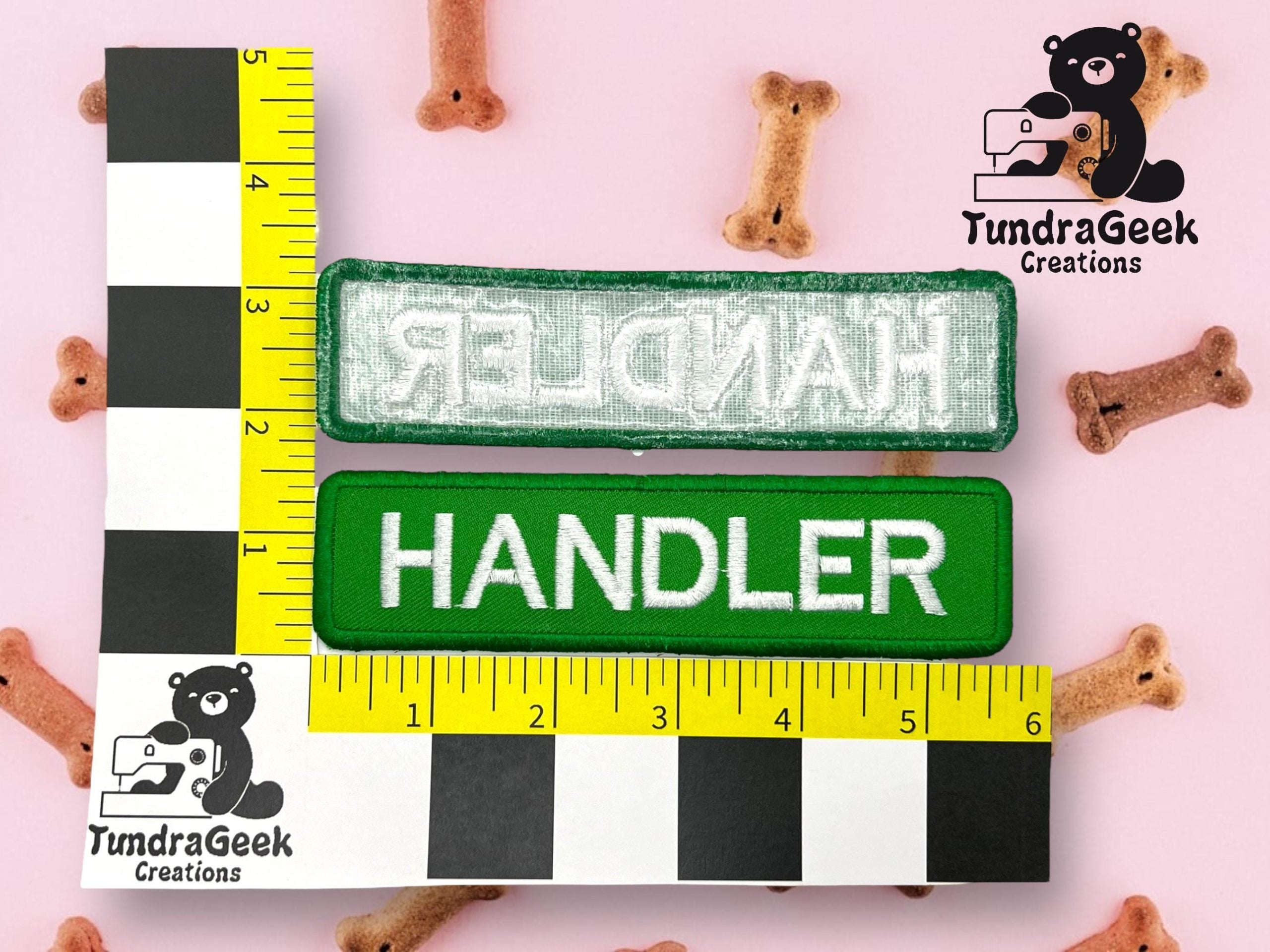 Two rectangular HANDLER patches, one facing up and one showing the back, are placed on a measurement grid with dog bone treats scattered on a pink background.