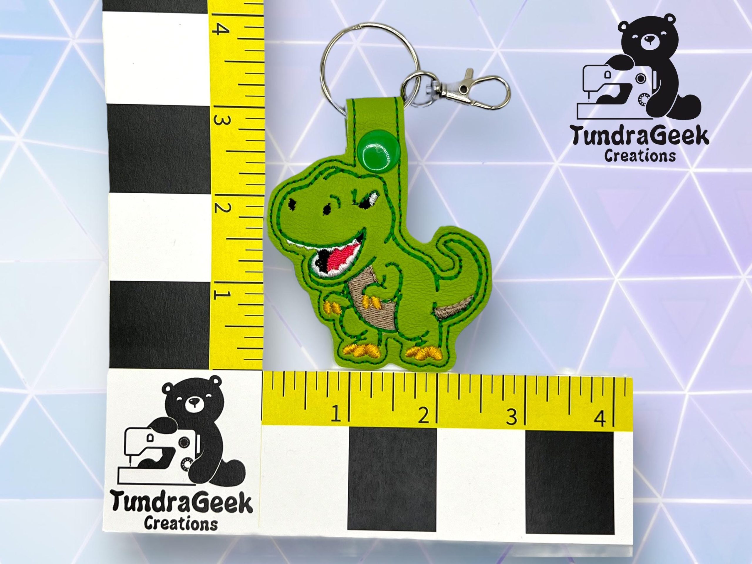 A green dinosaur keychain with a snap button lies on a measured grid. Logos reading TundraGeek Creations with a bear and sewing machine are visible in the corners.