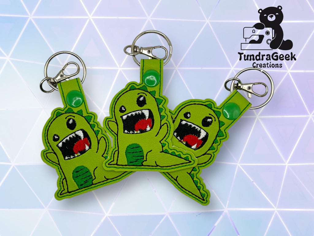 Three green dinosaur keychains with open mouths and stitched details are arranged on a geometric background; TundraGeek Creations logo is in the top right corner.