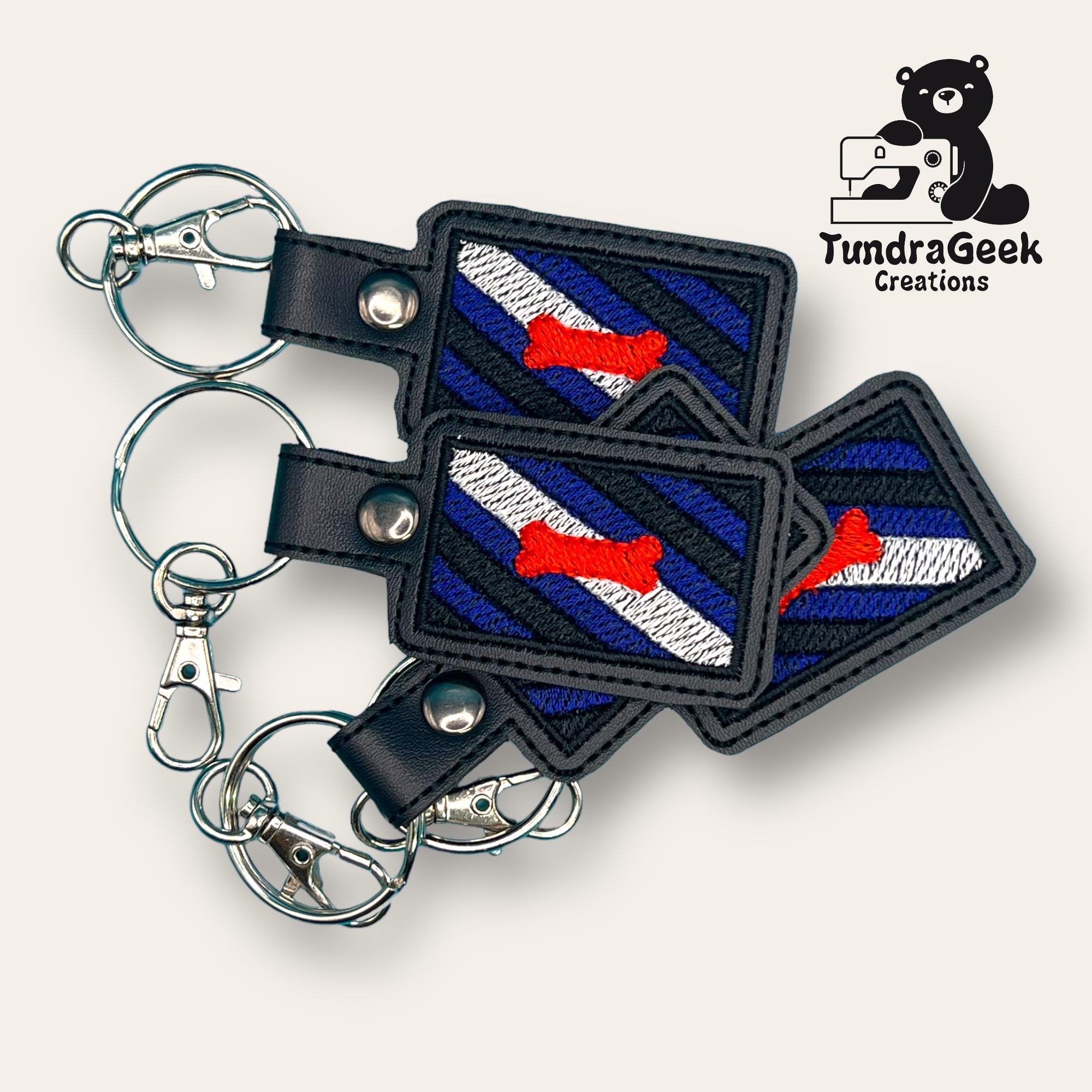 Three rectangular embroidered keychains with blue and black stripes, a white diagonal stripe, and a red bone design, displayed with metal key rings. TundraGeek Creations logo in the corner.