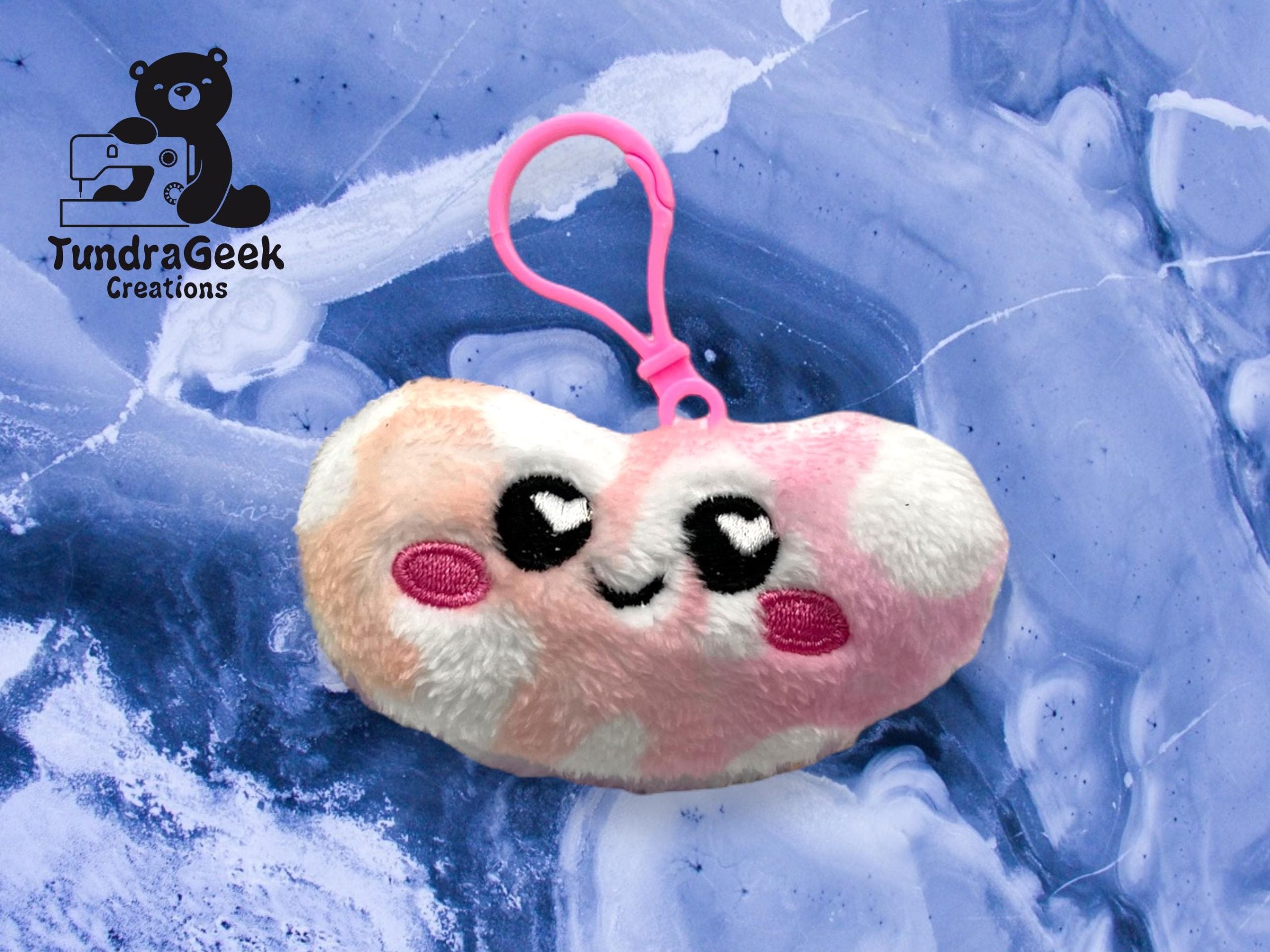 A plush toy keychain shaped like a bean with a smiling face, pink and white patches, on a blue marbled background. TundraGeek Creations logo appears in the upper left corner.