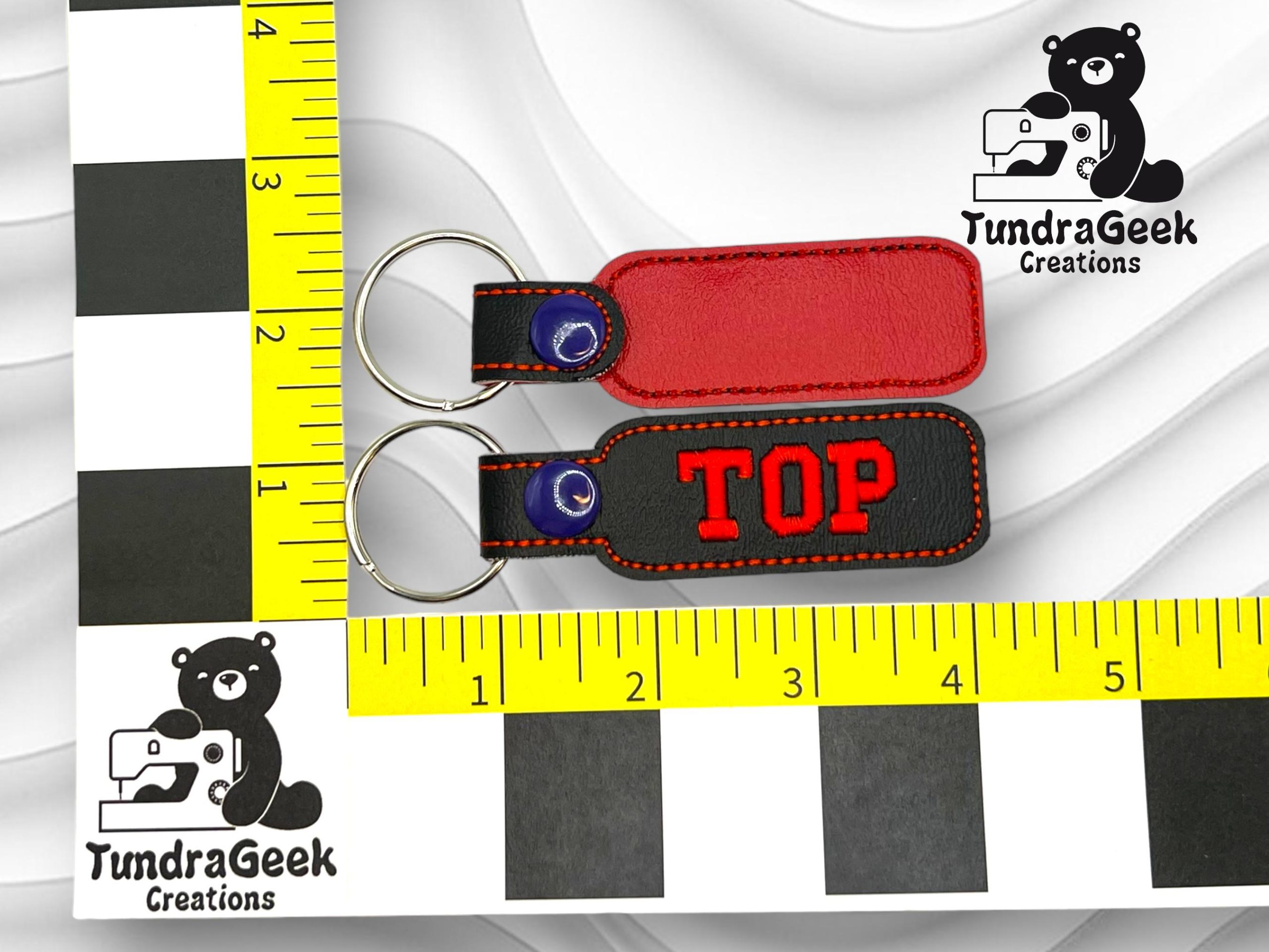 Two rectangular keychains, one red and one black with TOP in red letters, placed on a background with measuring rulers and TundraGeek Creations logos.