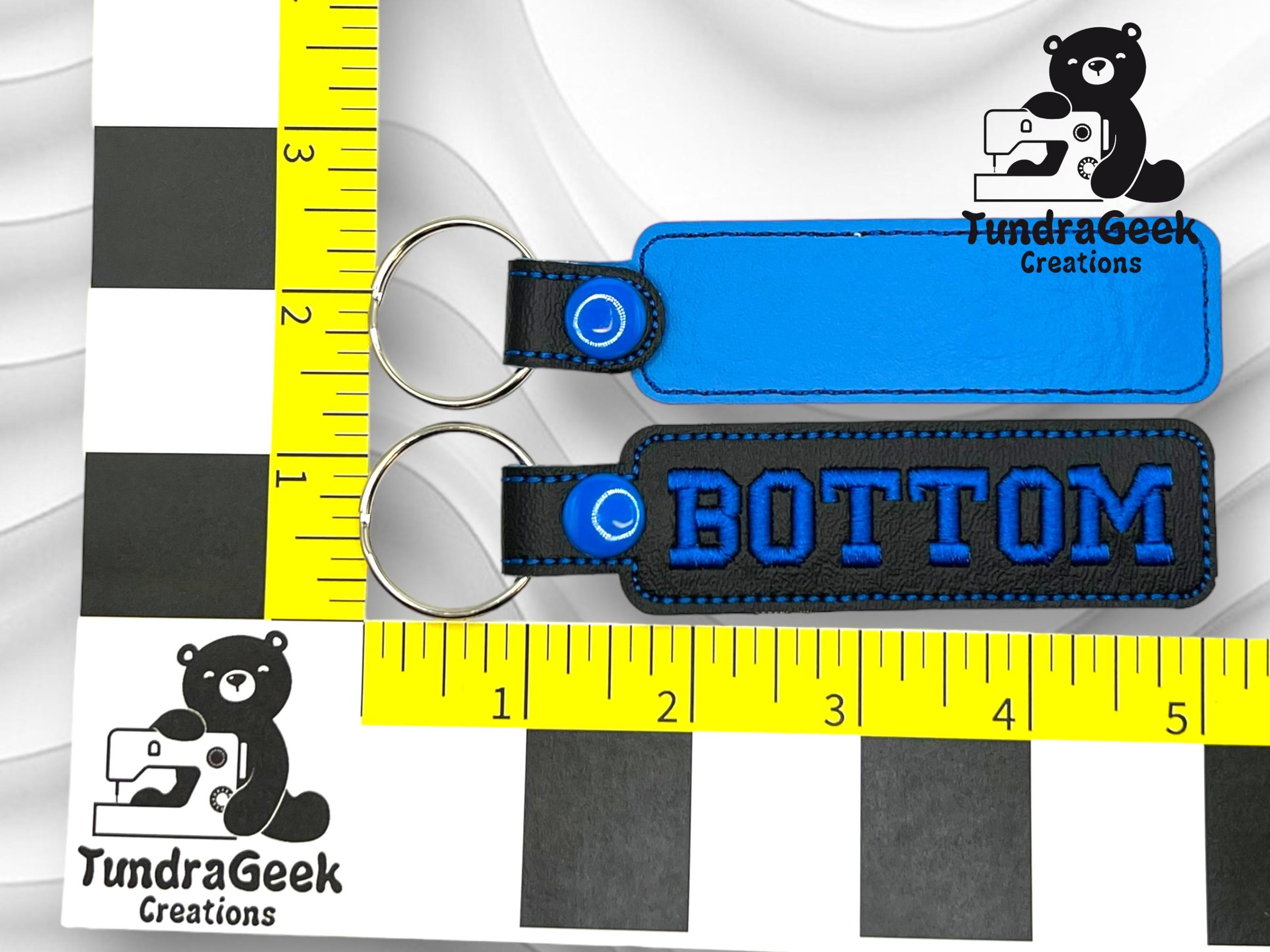 Two rectangular keychains are placed on a measurement guide. One is plain blue, the other is black with blue edges and the word BOTTOM. Both have metal key rings.