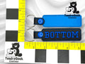 Two rectangular keychains on a measurement grid; one is plain blue, the other is black with blue trim and embroidered with the word BOTTOM. TundraGeek Creations logo in corners.
