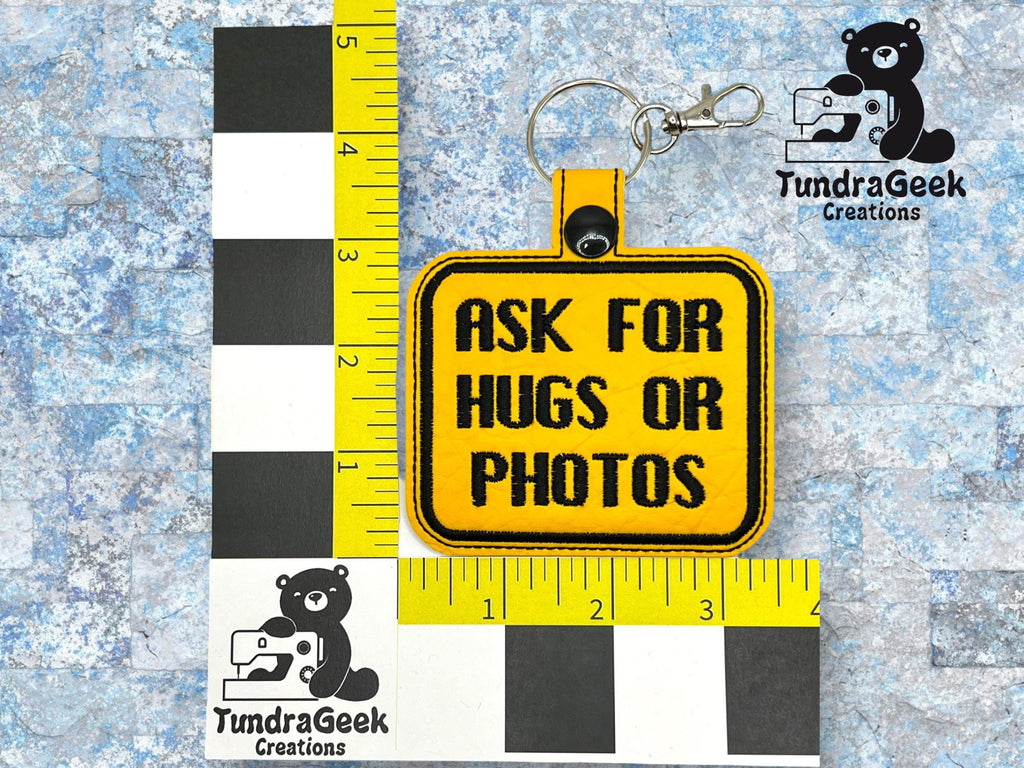 A yellow keychain with black text reading ASK FOR HUGS OR PHOTOS is placed on a grid with a TundraGeek Creations logo featuring a bear with a sewing machine.