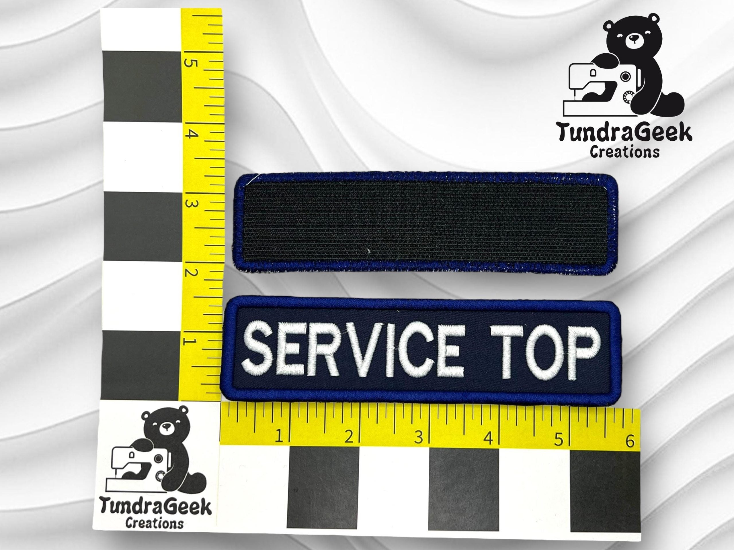 Two rectangular patches are placed on a ruler; one is blank and black, the other is blue with white text reading SERVICE TOP. TundraGeek Creations logo appears in two corners.