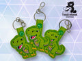 Three green dinosaur keychains with smiling faces displayed on a geometric background, with the TundraGeek Creations logo in the upper right corner.