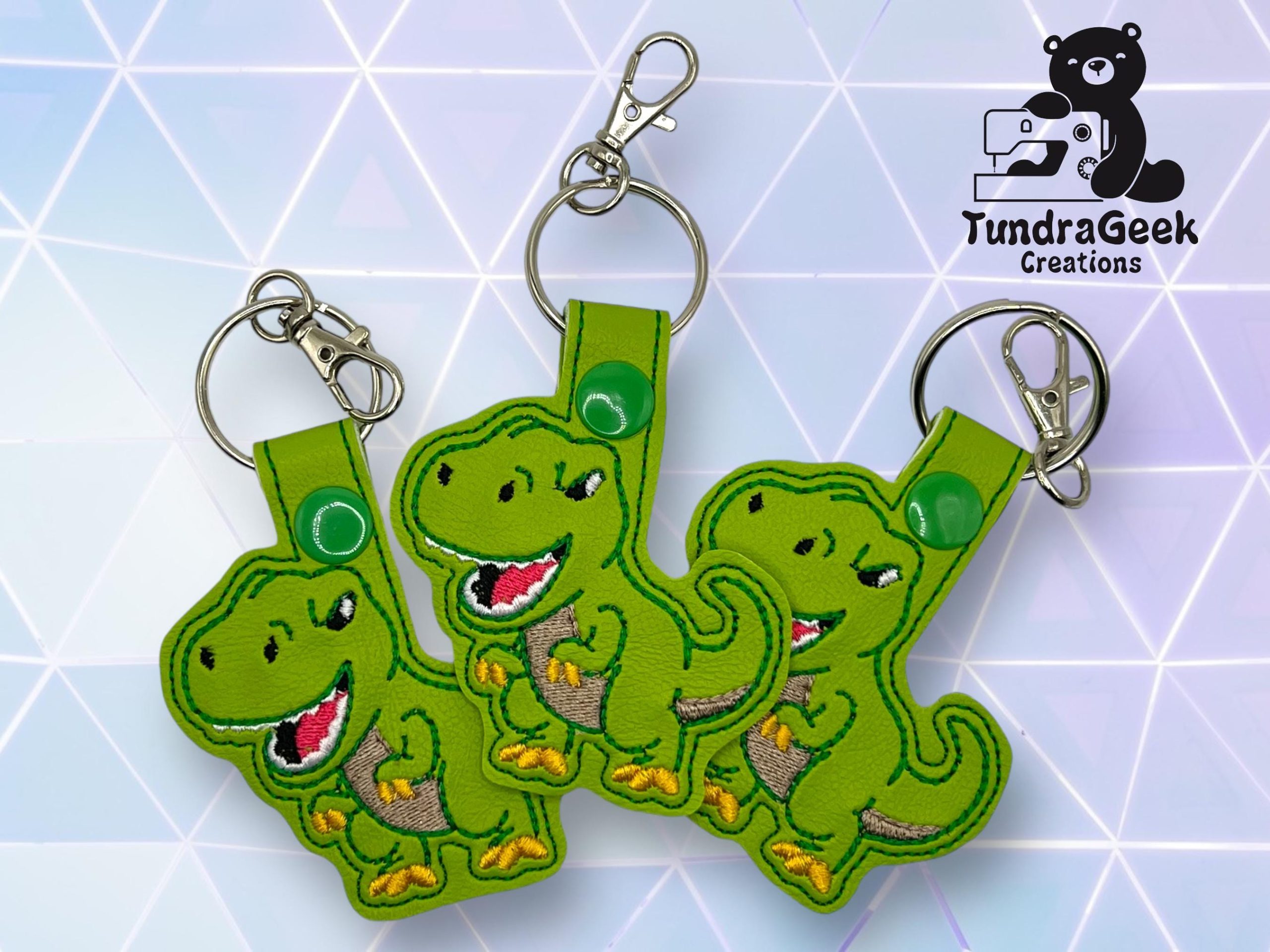 Three green dinosaur keychains with smiling faces displayed on a geometric background, with the TundraGeek Creations logo in the upper right corner.