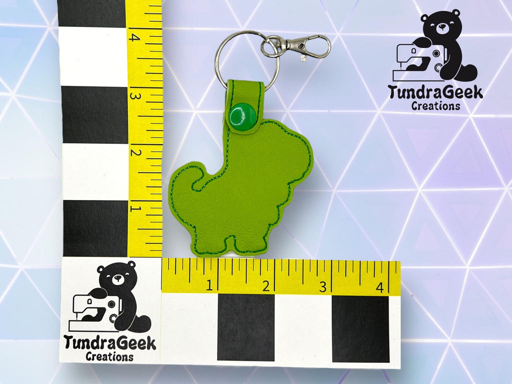 A green dinosaur-shaped keychain is placed next to a ruler measuring about 4 inches. The TundraGeek Creations logo appears twice in the image.