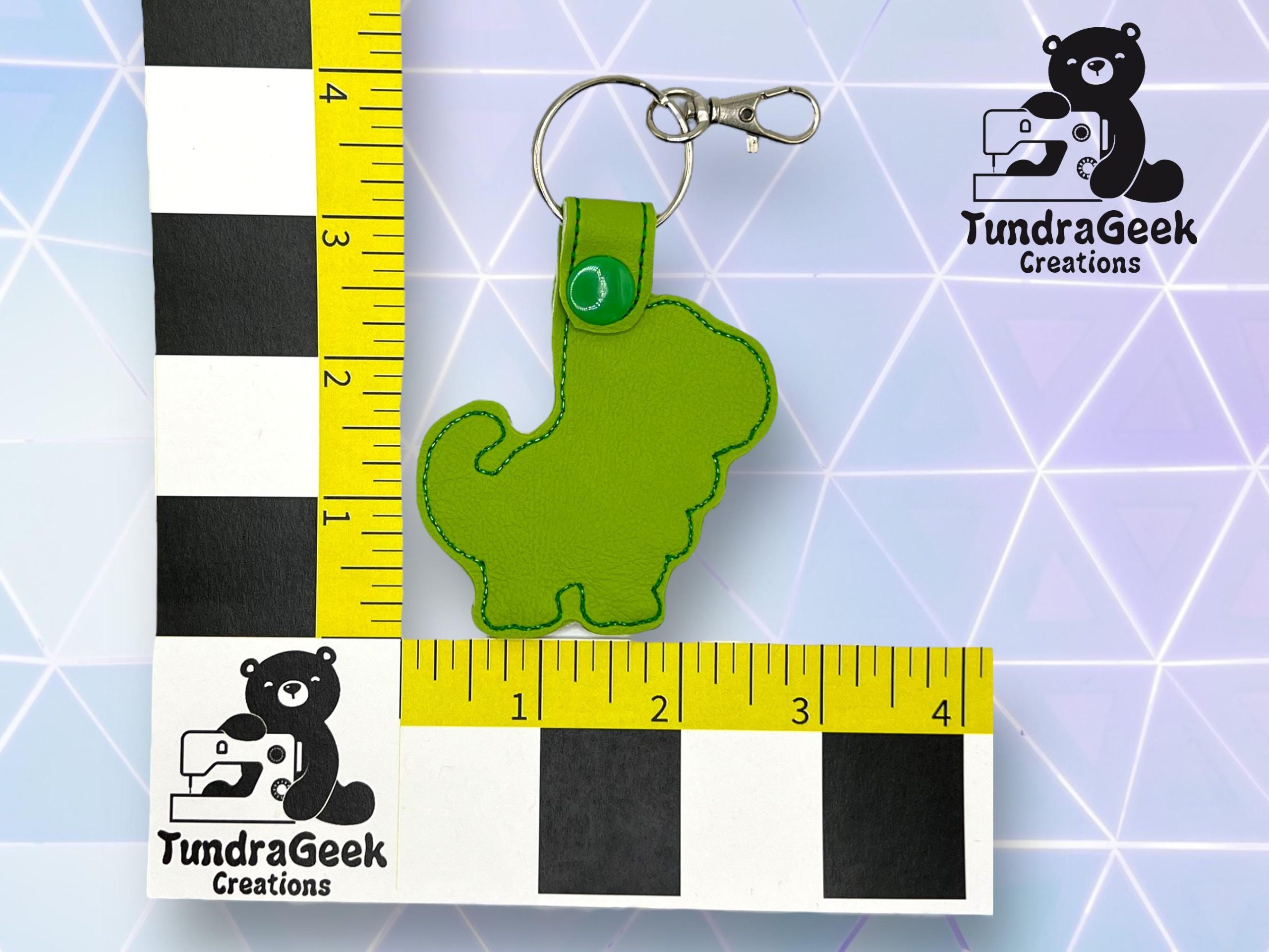 A green dinosaur-shaped keychain is placed next to a ruler measuring about 4 inches. The TundraGeek Creations logo appears twice in the image.