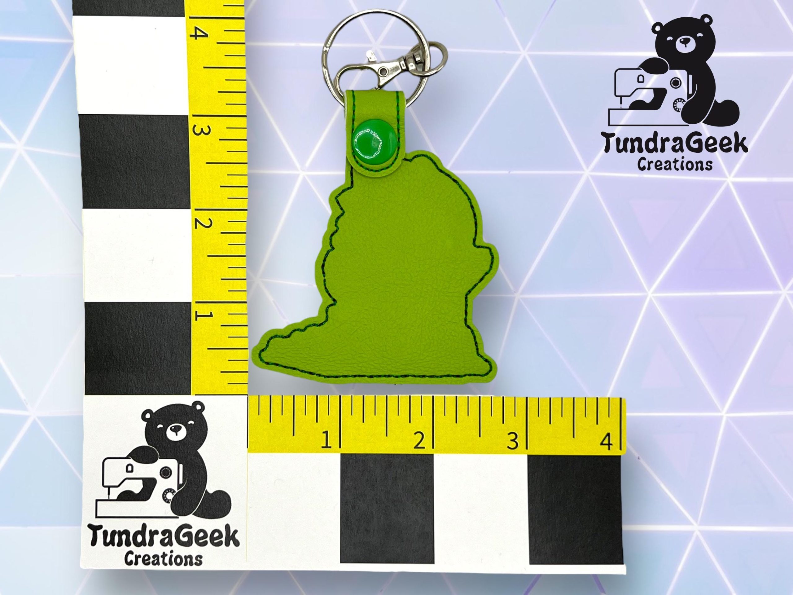 A green keychain shaped like a head, shown next to two measuring rulers for scale on a geometric background with the TundraGeek Creations logo.