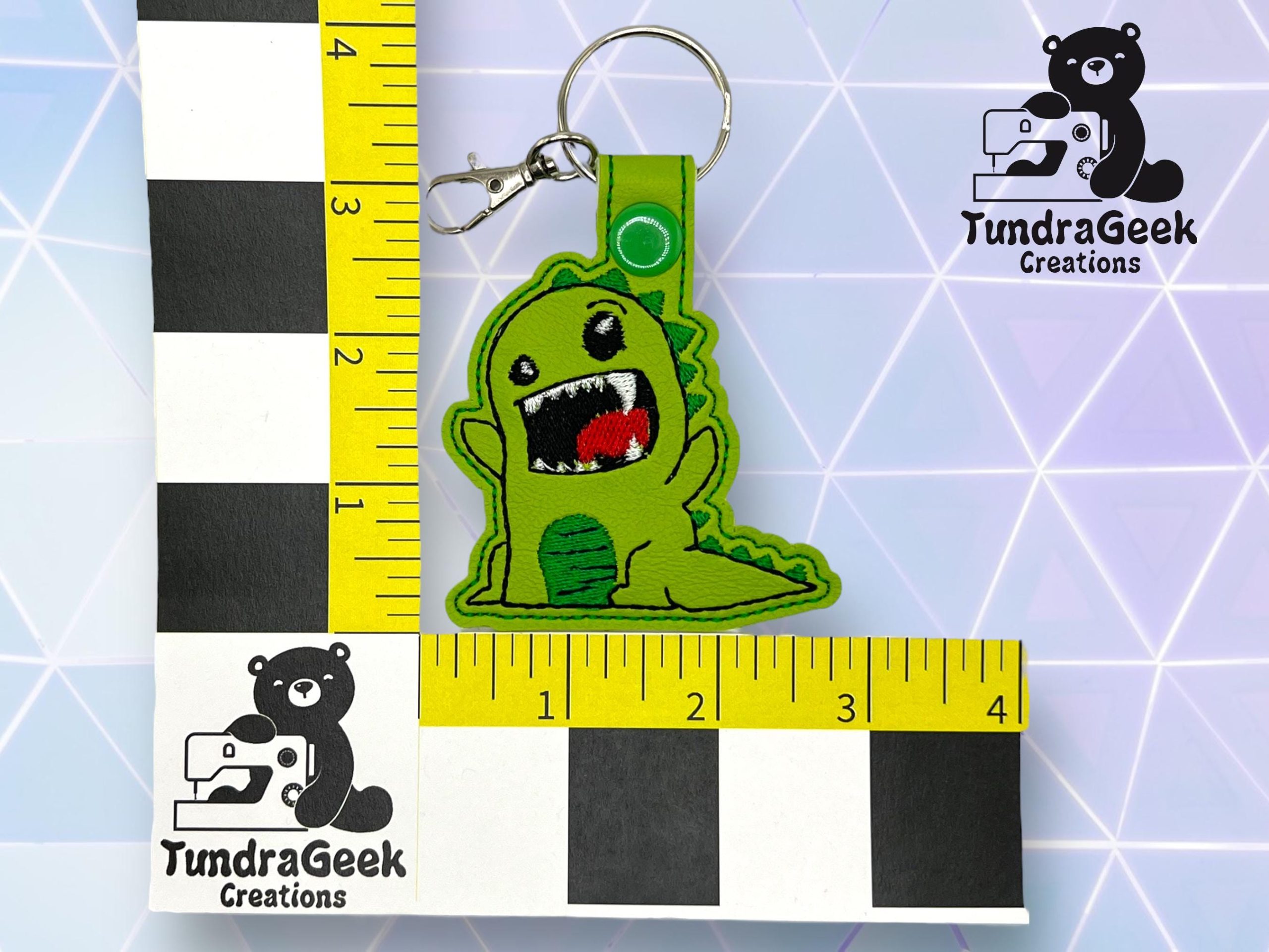 A green dinosaur keychain with an open mouth is placed on a checkered measurement grid beside TundraGeek Creations logos and yellow measuring tape.