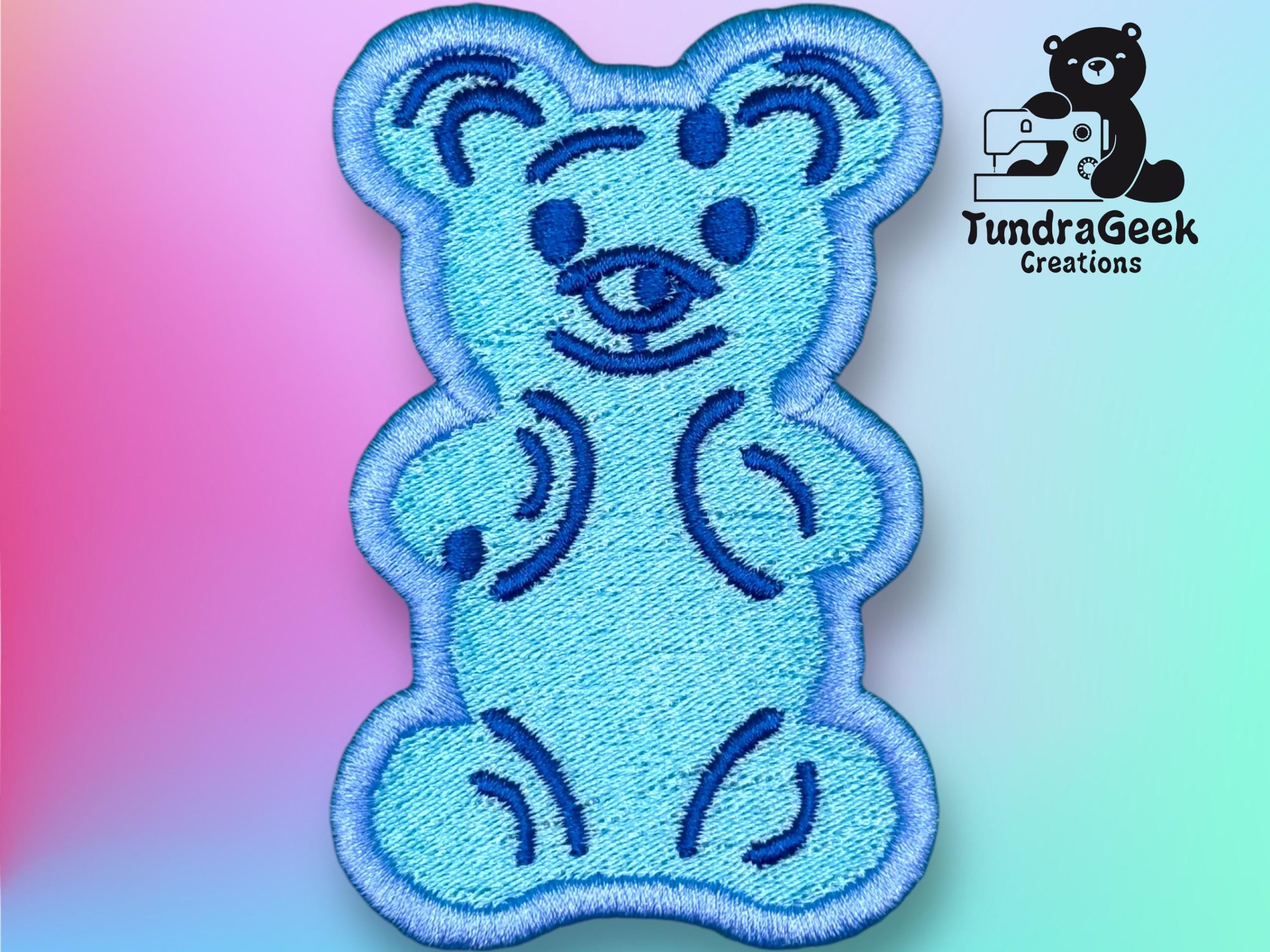 Blue embroidered bear patch with dark blue outline and details, shown on a pastel gradient background. TundraGeek Creations logo appears in the top right corner.