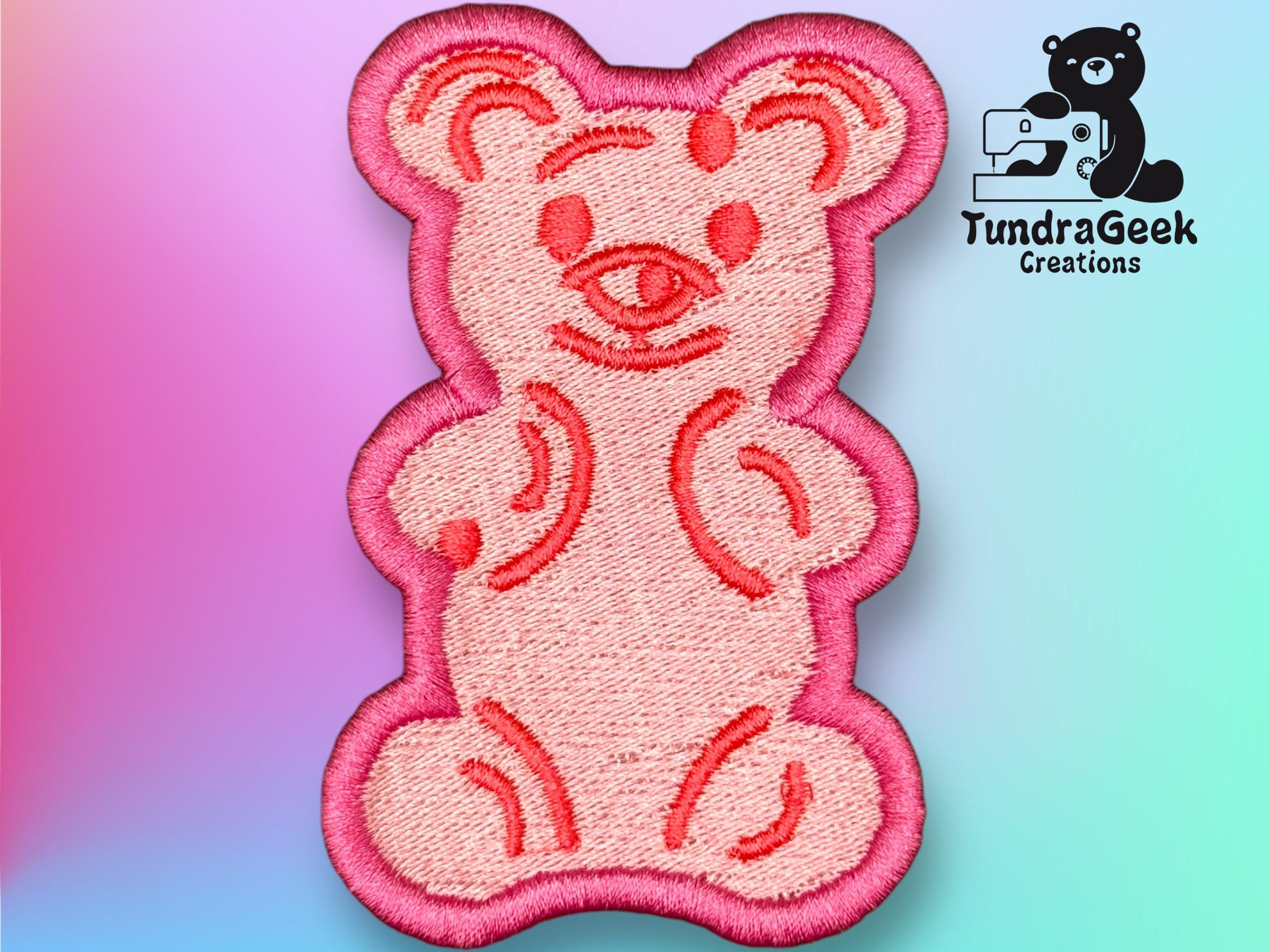 Pink embroidered bear patch with simple facial and body details, shown on a pastel gradient background. TundraGeek Creations logo with a bear and sewing machine in the top right corner.