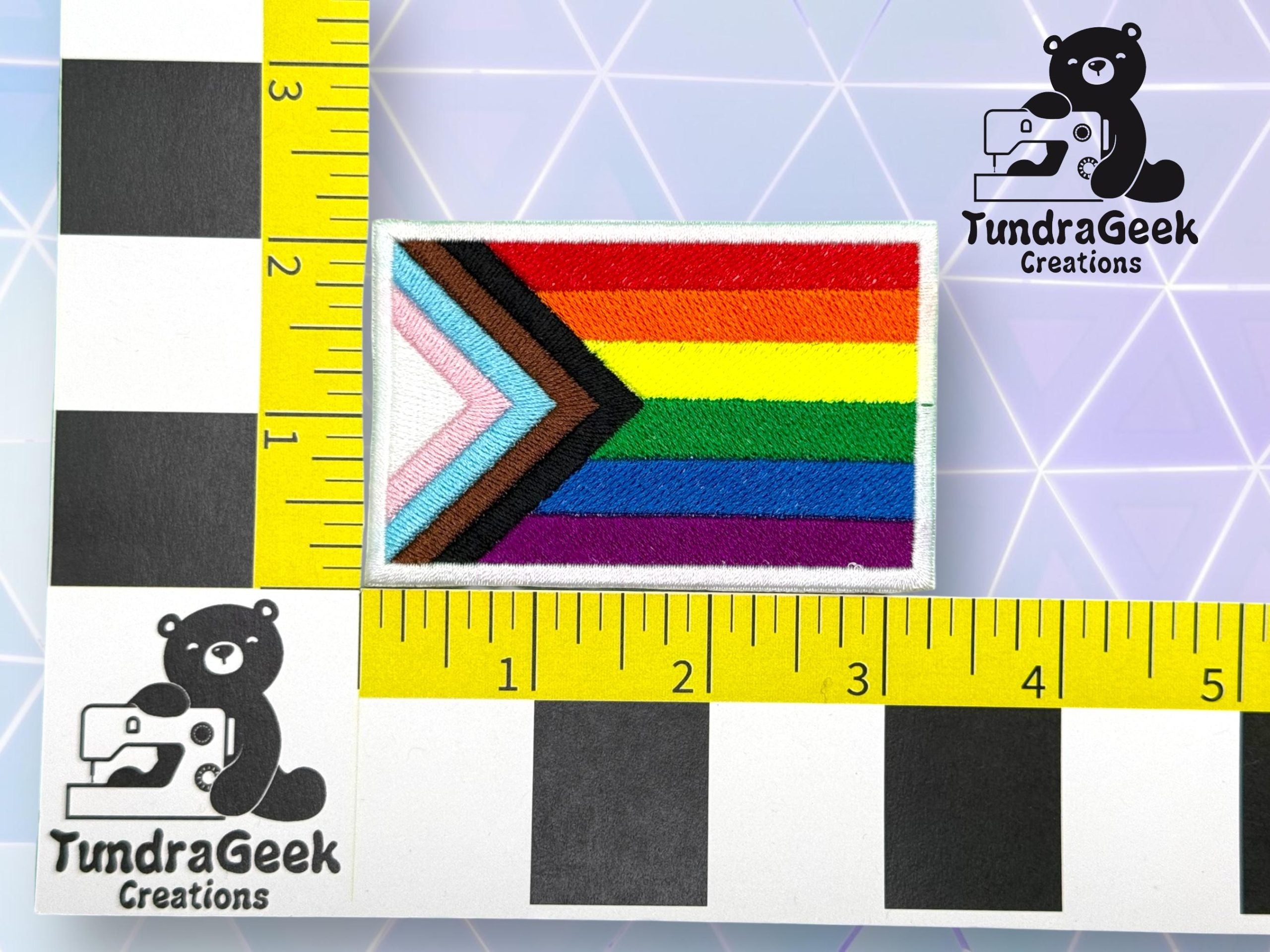A rectangular Progress Pride flag patch is measured against a ruler, showing a size of about 2 by 3 inches. The logo TundraGeek Creations with a bear is visible in two corners.