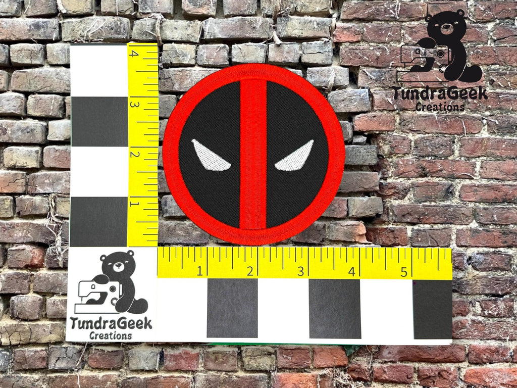 A round patch with stylized white eyes on a black and red background is placed on a measurement grid against a brick wall. The patch measures roughly 3 inches in diameter.