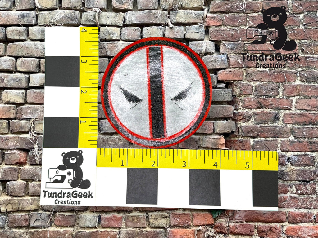 A circular embroidered patch with black and red details is measured against a ruler, placed on a brick background. TundraGeek Creations logo appears twice in the image.