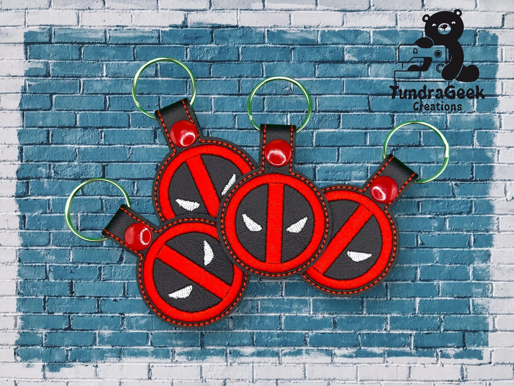 Four red and black keychains with a masked face design are displayed on a blue brick background. The TundraGeek Creations logo is in the top right corner.