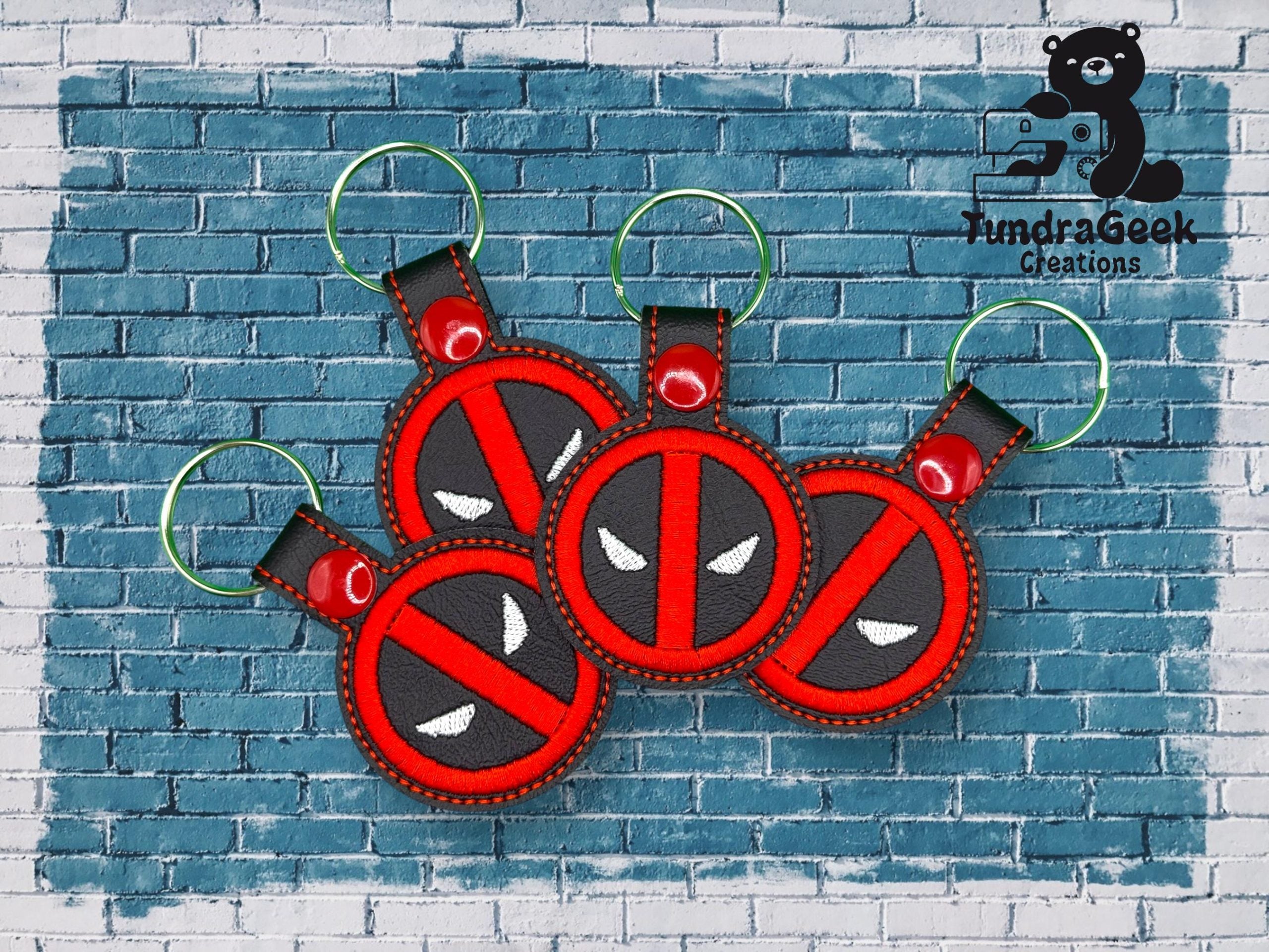 Four red and black keychains with a masked face design are displayed on a blue brick background. The TundraGeek Creations logo is in the top right corner.