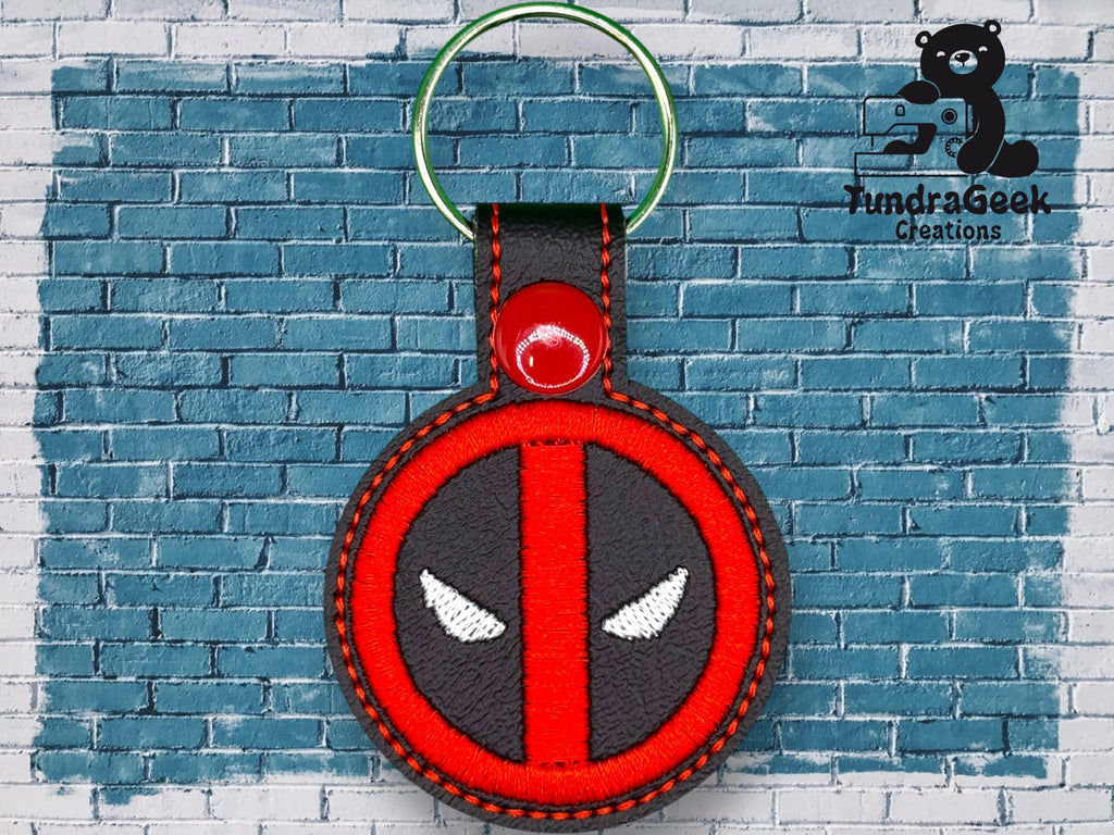 A round keychain with a red and black design resembling Deadpool’s logo is displayed against a blue brick wall with the TundraGeek Creations logo in the top right corner.