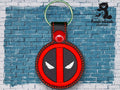 A round keychain with a red and black design resembling Deadpool’s logo is displayed against a blue brick wall with the TundraGeek Creations logo in the top right corner.
