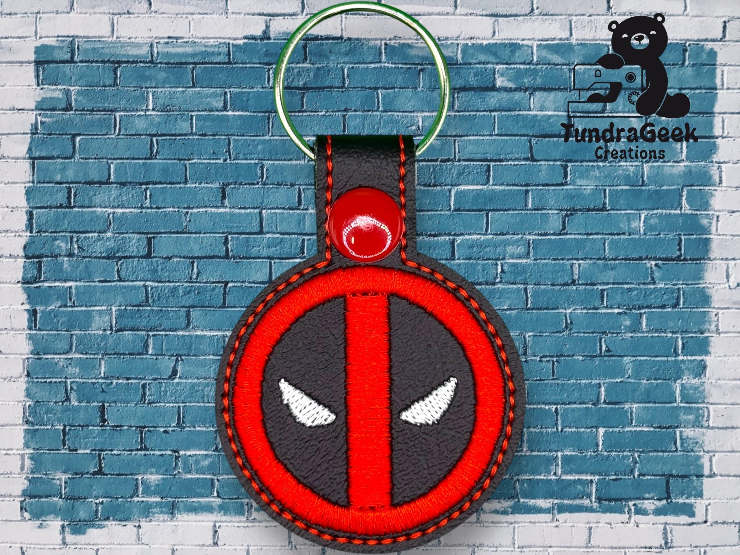 A round keychain with a red and black design resembling Deadpool’s logo is displayed against a blue brick wall with the TundraGeek Creations logo in the top right corner.