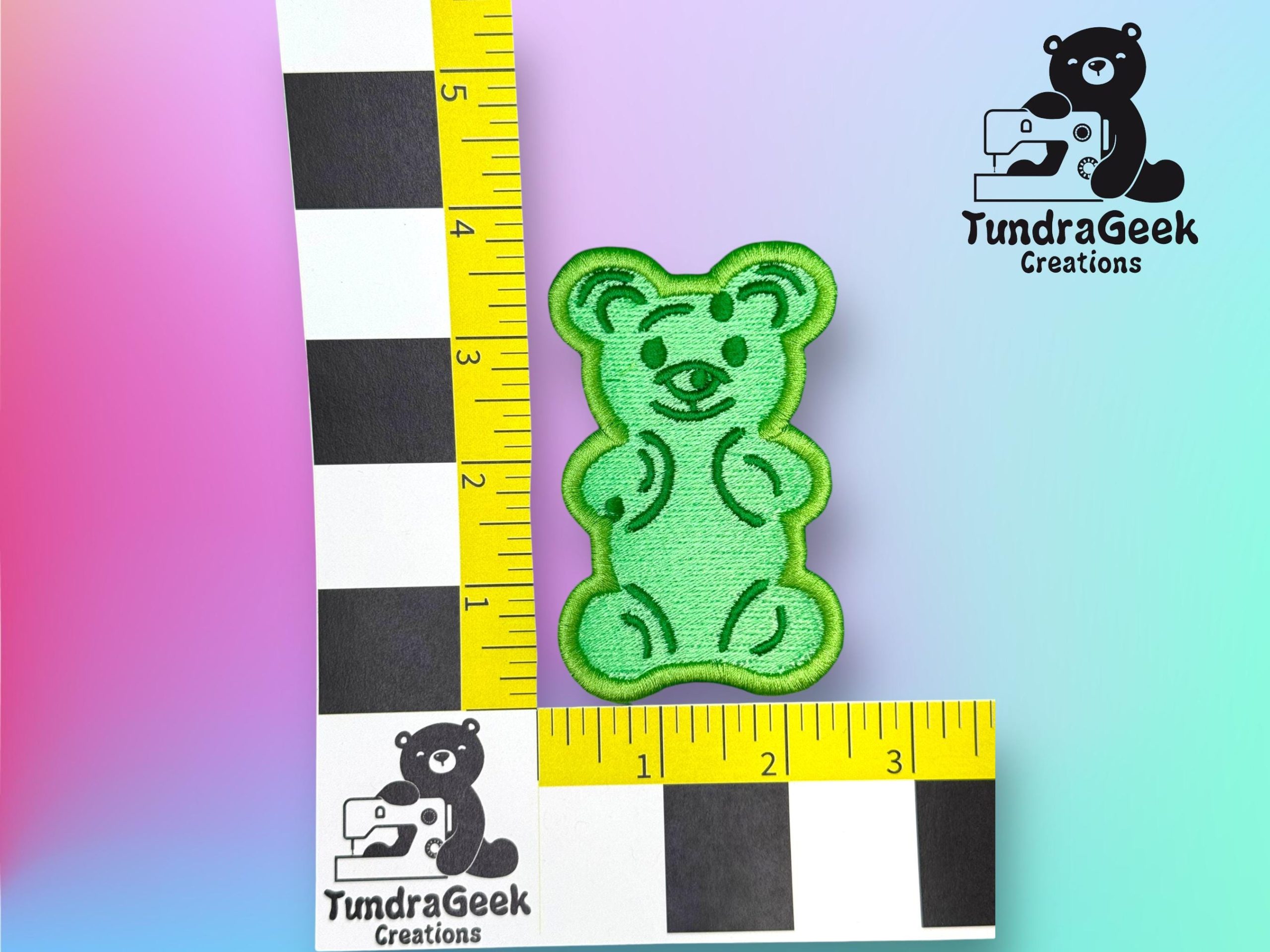 A green embroidered bear patch is placed next to a yellow and black ruler for scale, with the TundraGeek Creations logo in two corners on a pastel gradient background.