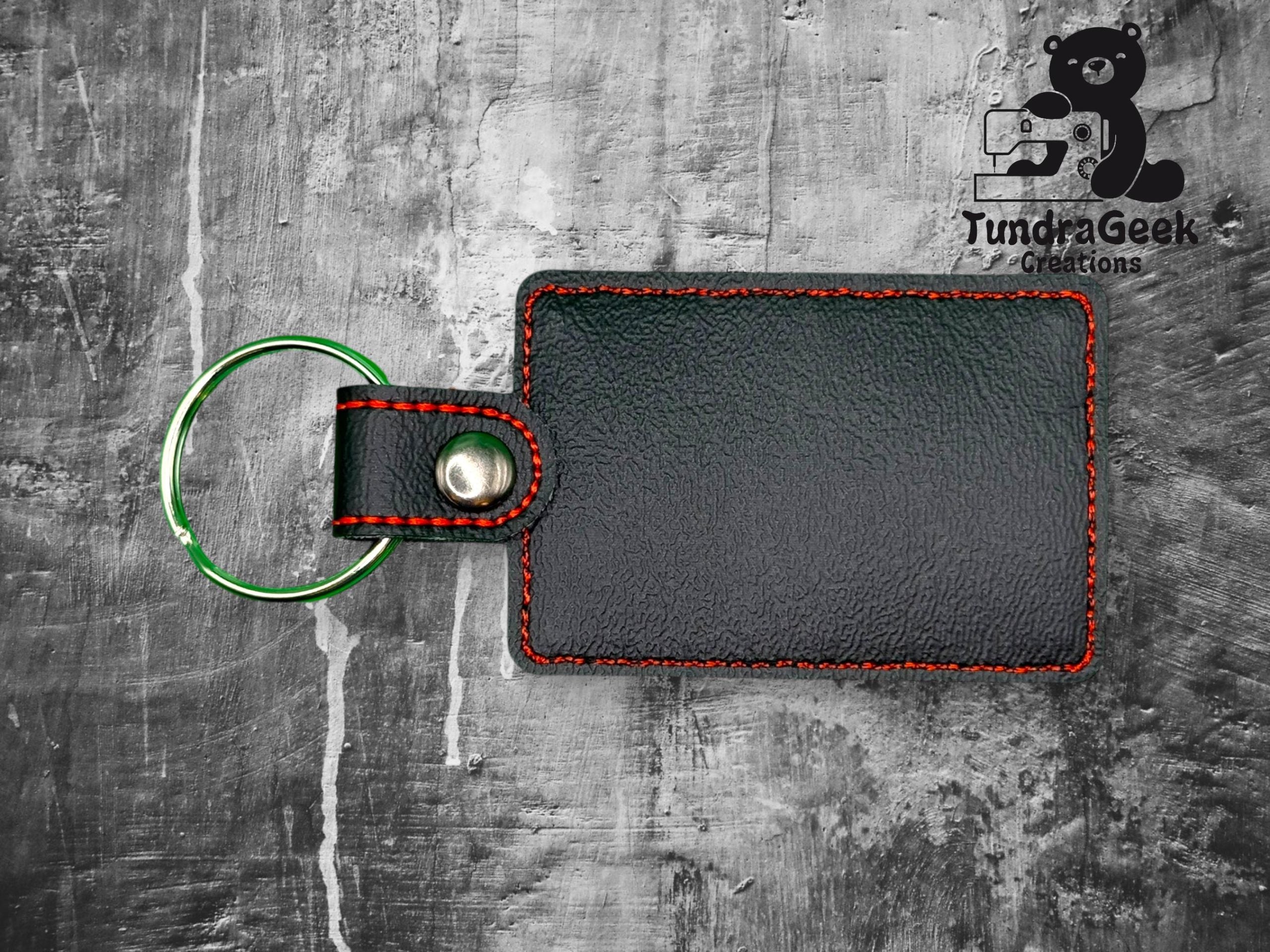 Rectangular black leather keychain with red stitching and a silver key ring on a textured gray surface. TundraGeek Creations logo is in the top right corner.