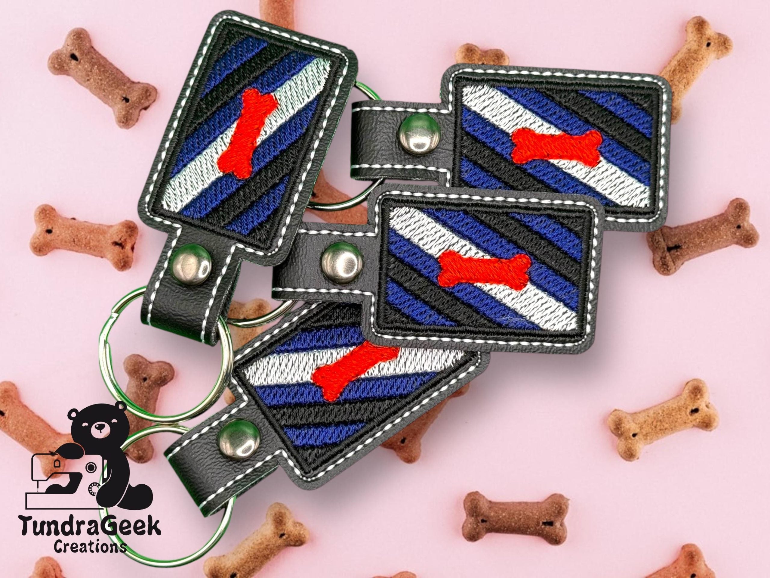 Four rectangular keychains with a blue, black, white, and red embroidered design are displayed on a pink surface scattered with dog bone-shaped treats. TundraGeek Creations logo is present.