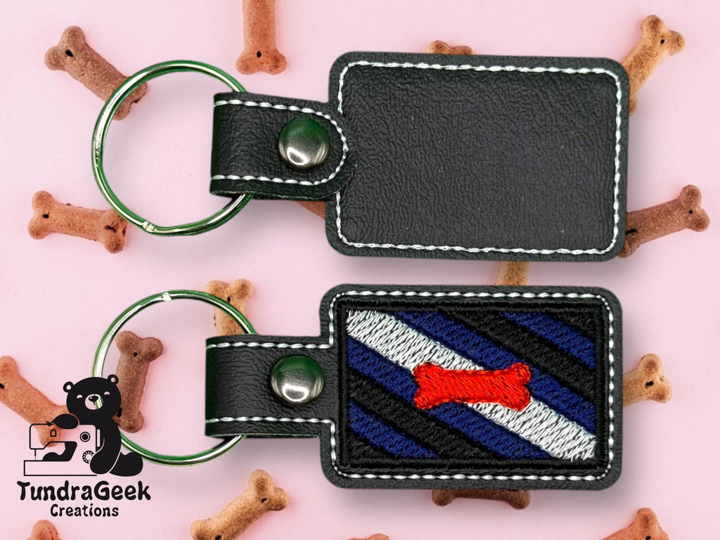 Two black leather keychains with white stitching are shown; one features embroidery of a red dog bone on a blue and black striped background. Scattered dog biscuits and a TundraGeek Creations logo appear.