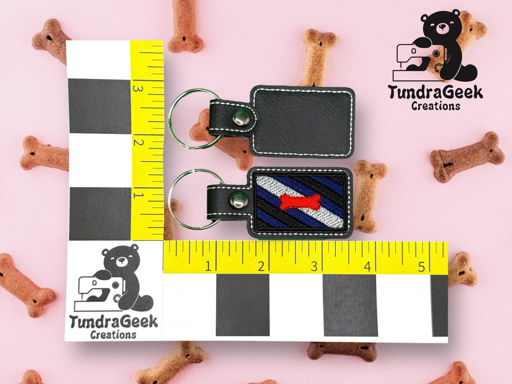 Two black rectangular keychains, one blank and one embroidered with a red dog bone, are placed next to a ruler for scale. Dog treats and TundraGeek Creations logos appear in the background.